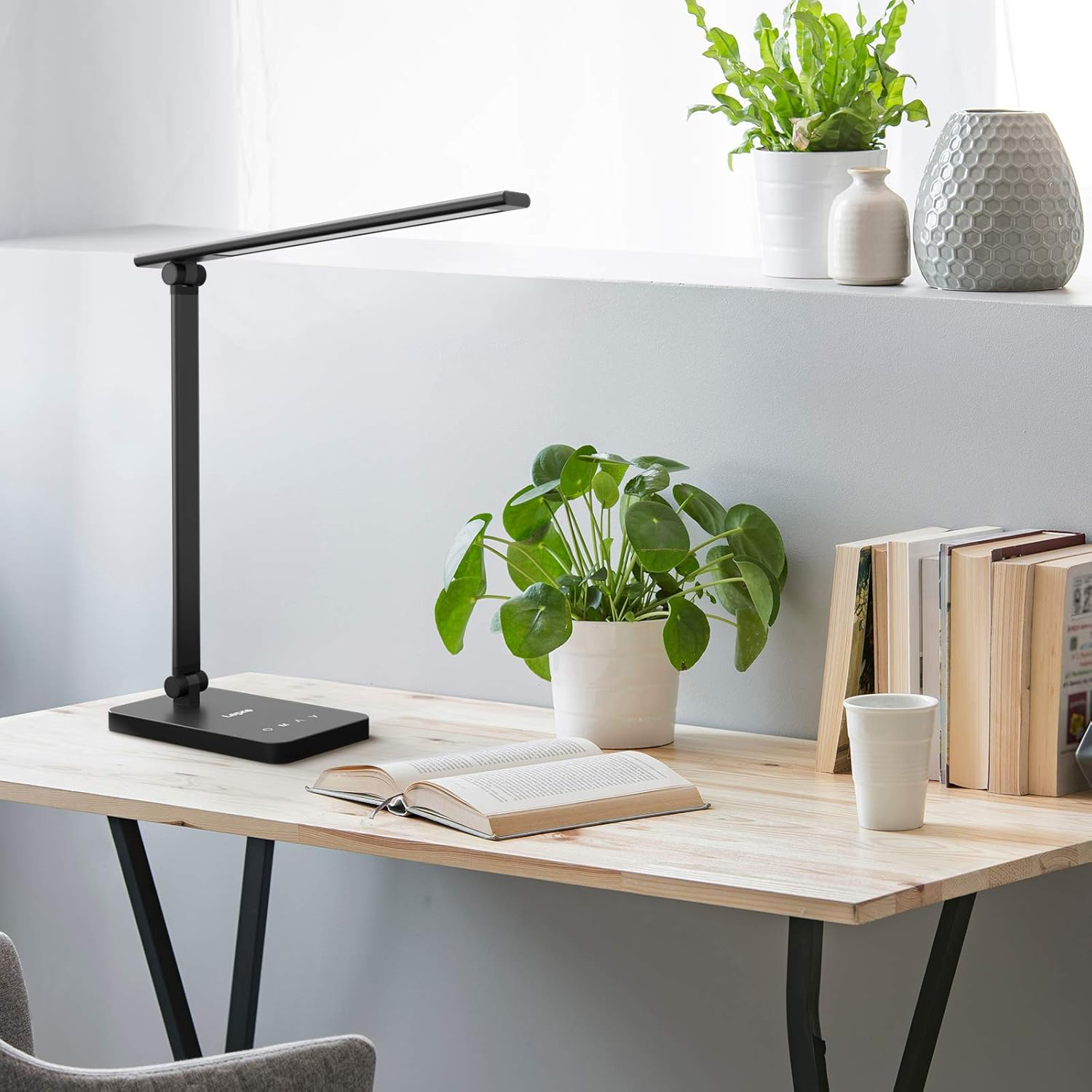 Schwarze Schreibtischlampe, offenes Buch, Pflanzen und Kaffeetasse auf einem Holzschreibtisch am Fenster.