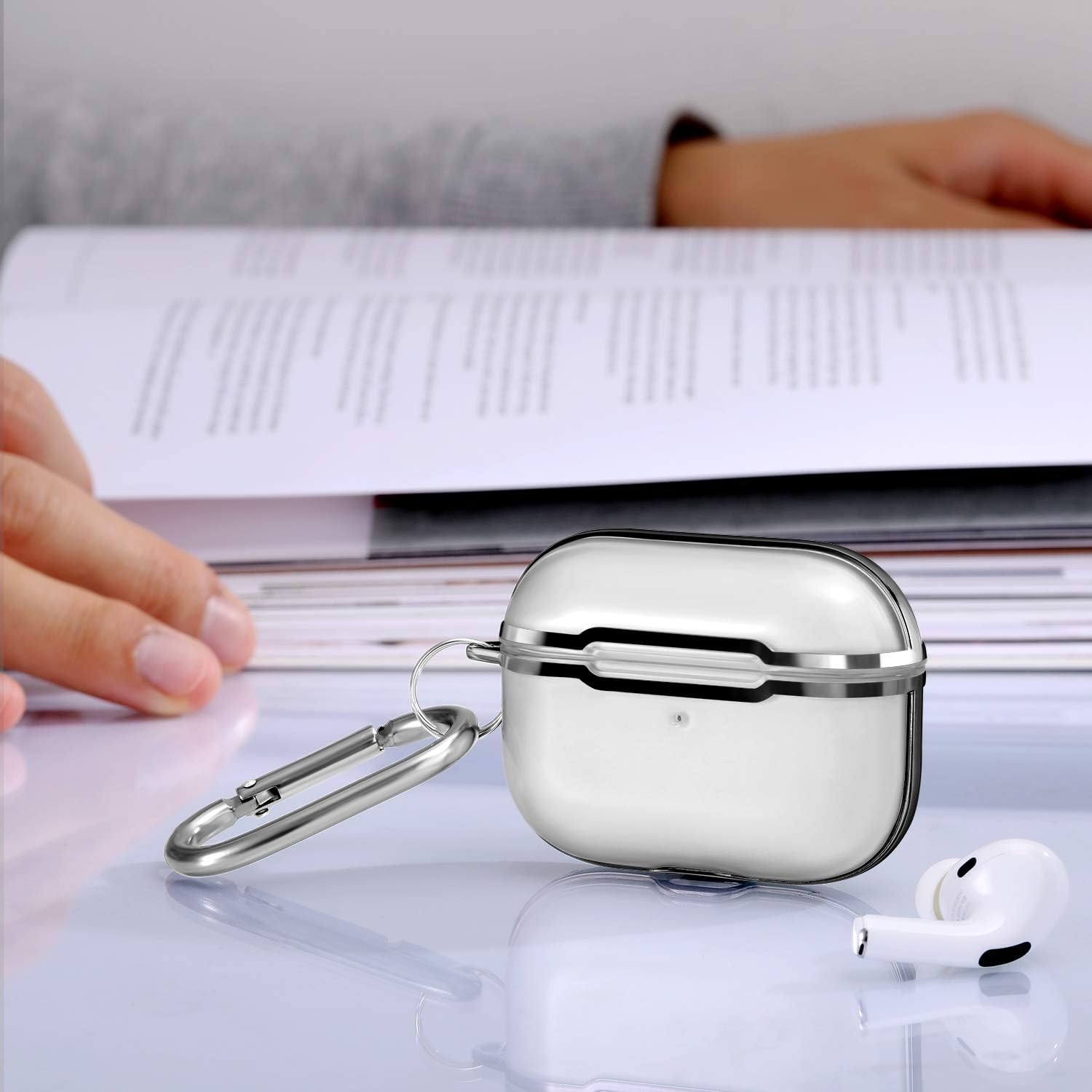 Weiße AirPods in einem weißen Etui mit einem silbernen Karabiner, auf einem Tisch mit einem verschwommenen Buch.