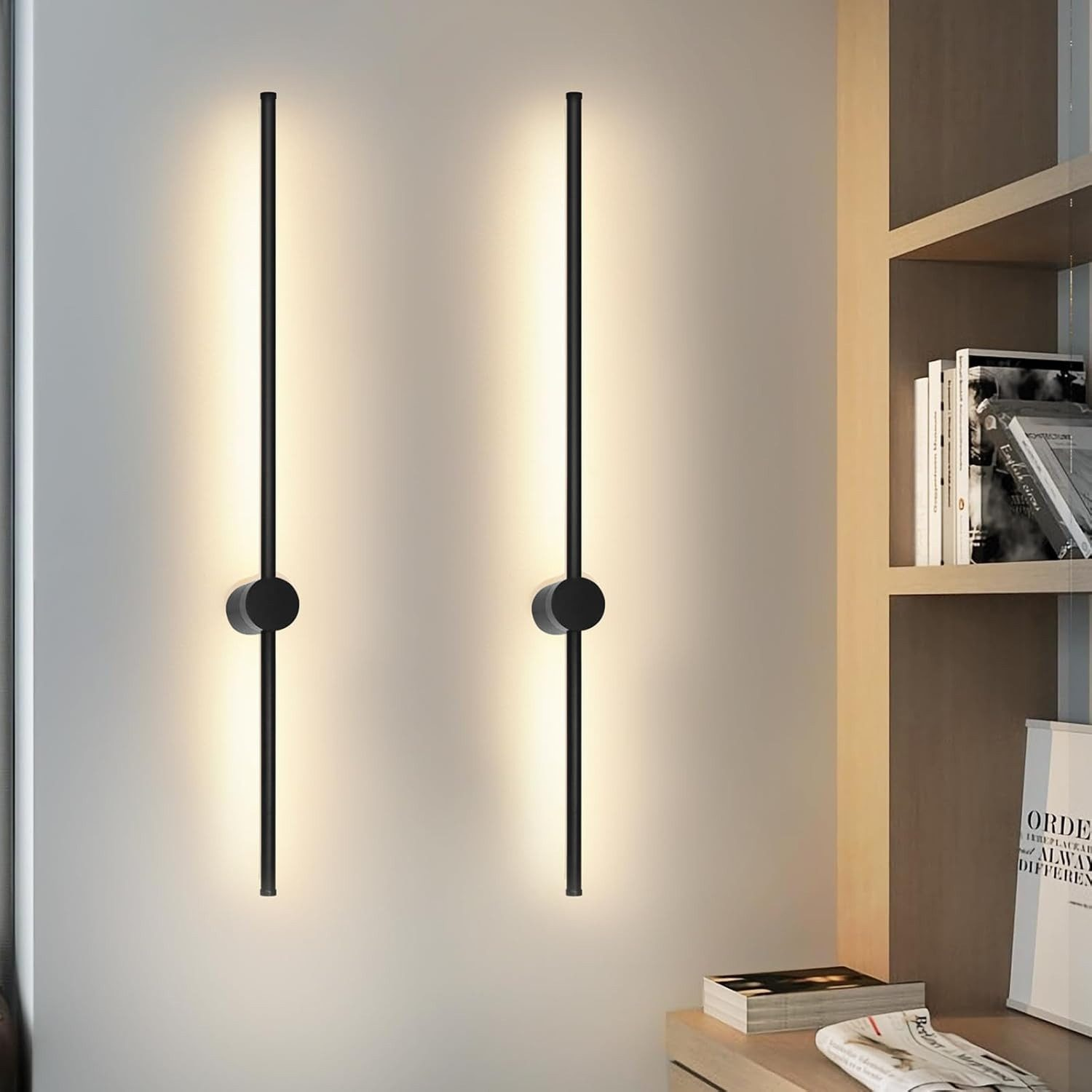 Due luci a LED nere verticali montate su una parete bianca, con una libreria visibile.