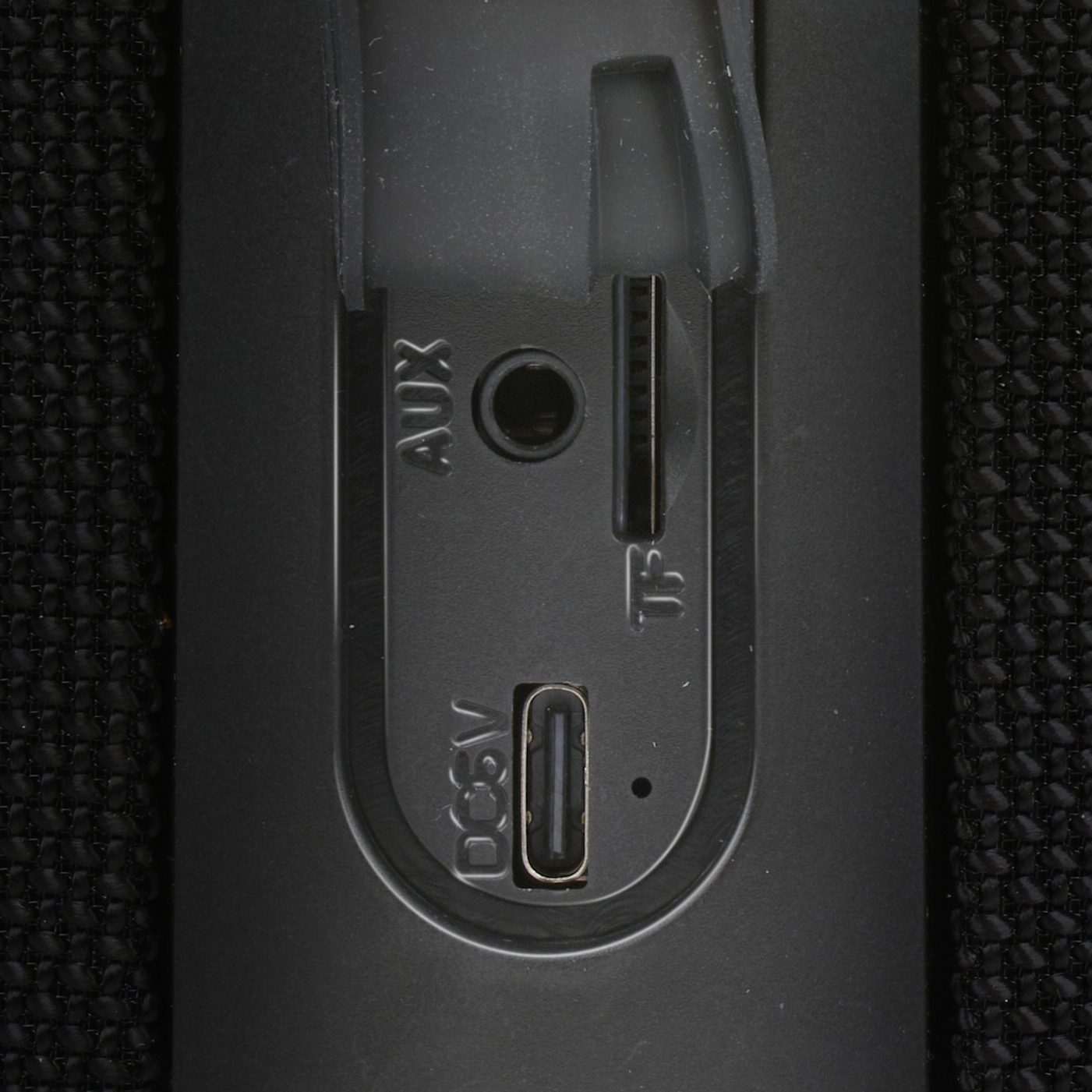 Close-up van luidsprekerpoorten: DC 5V, AUX, TF en een USB-poort, op een zwarte textuur.
