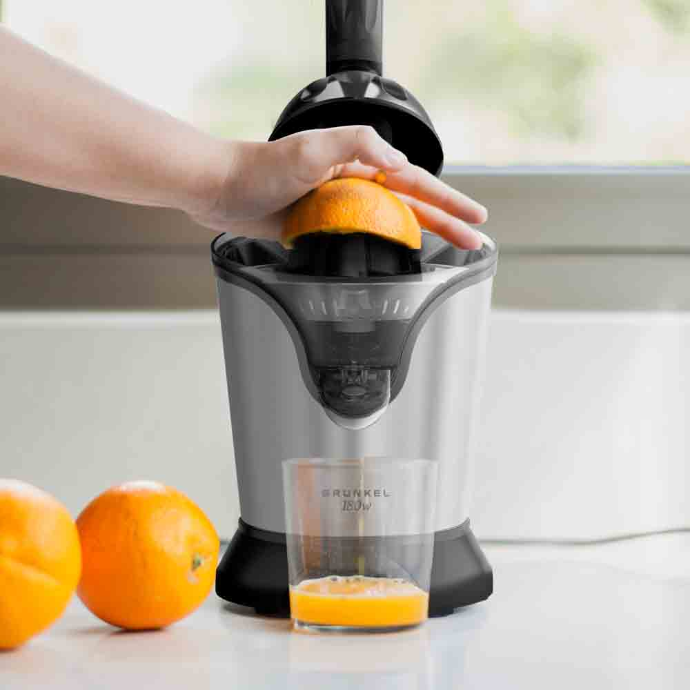 Persona che sprema un'arancia. Spremiagrumi argentato, bicchiere di succo, arance sul bancone. Cucina luminosa.