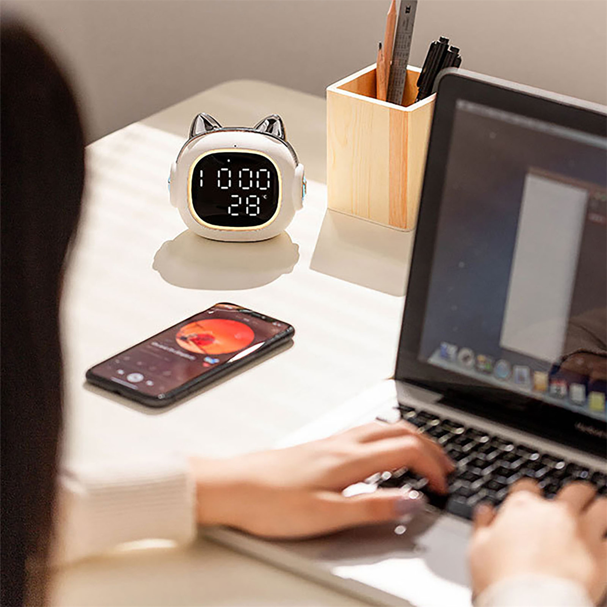 Eine Uhr mit Katzenohren, ein Telefon und ein Laptop auf einem Schreibtisch. Eine Person tippt auf dem Laptop.