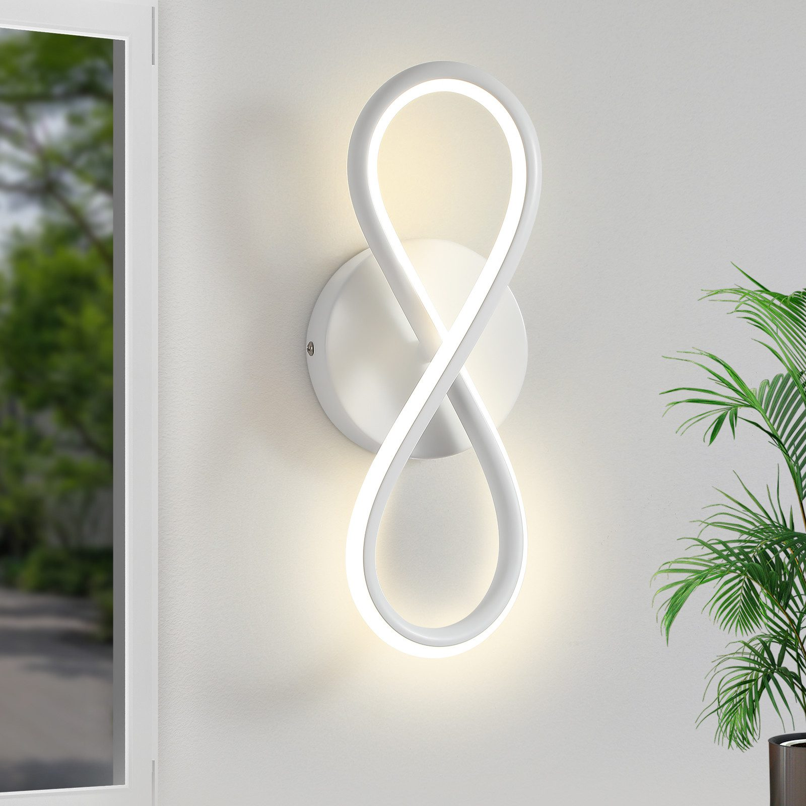 Una lampada da parete bianca a forma di infinito su un muro bianco. C'è una pianta verde a destra.