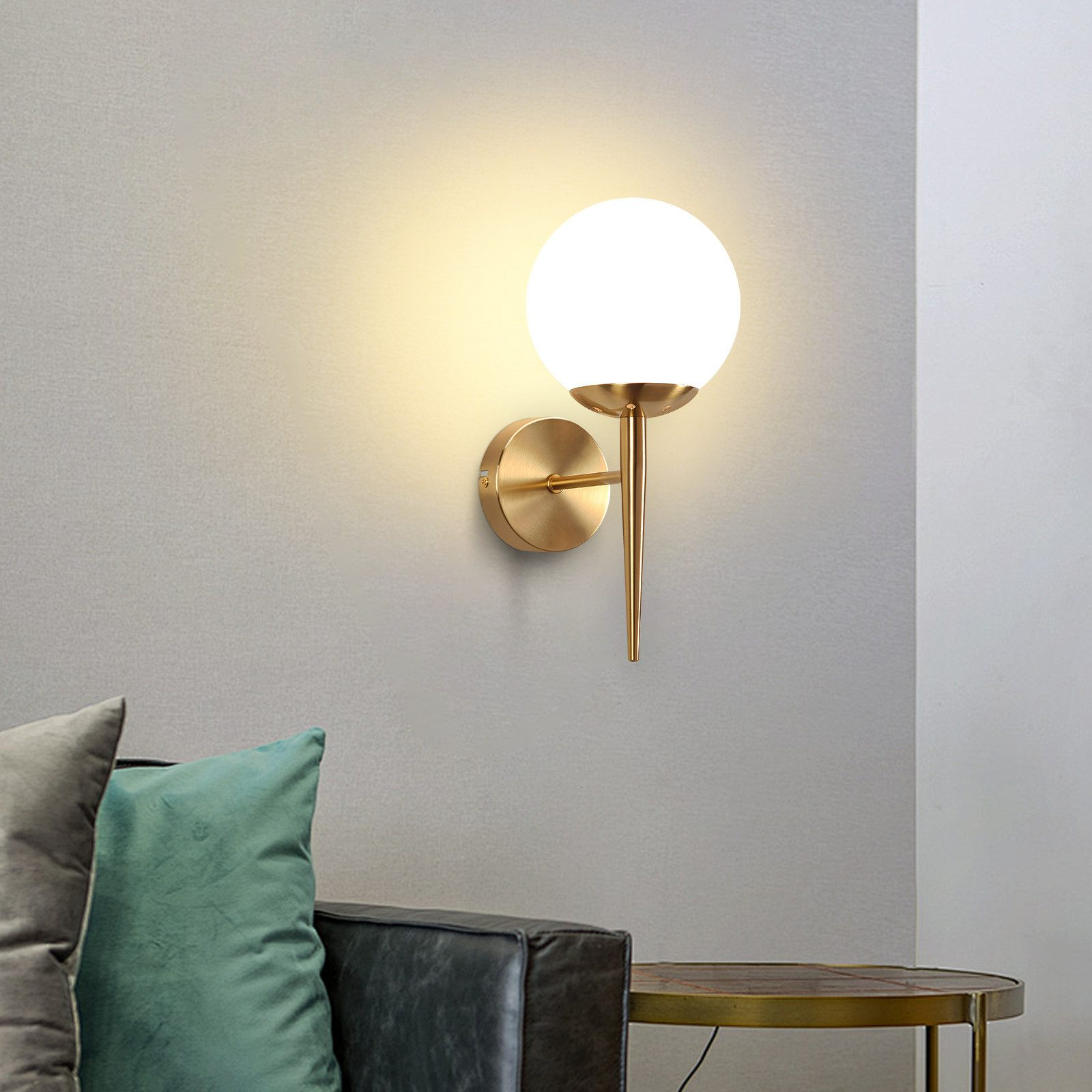 Lampada dorata montata a parete con paralume a globo bianco. Cuscino verde e divano nero sullo sfondo.