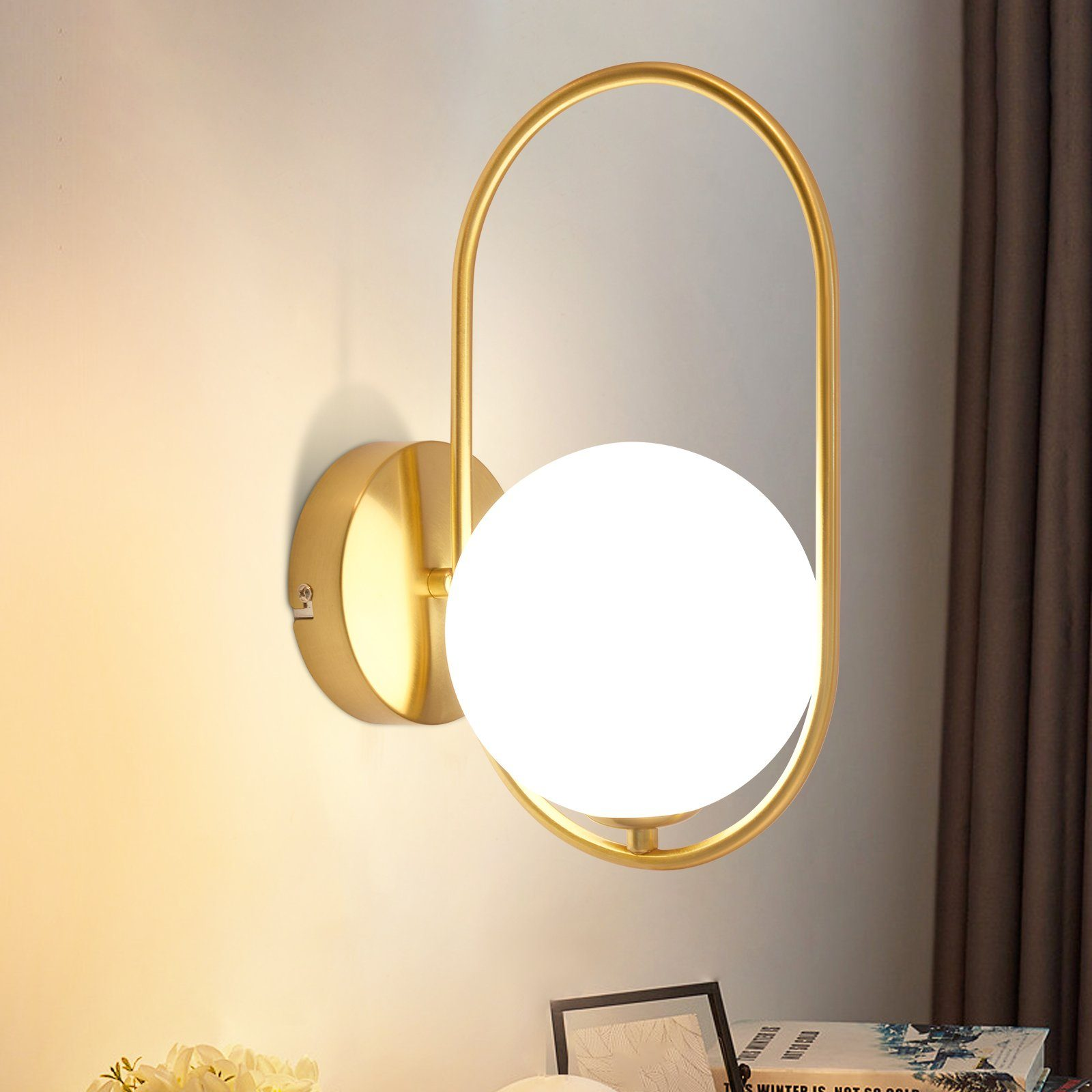Lampada da parete dorata con una luce a globo bianca, montata su una parete. Un libro e oggetti sotto.