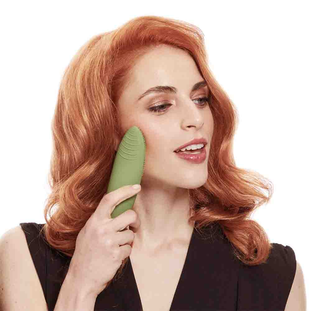Una mujer con cabello rojo usa un cepillo de limpieza facial verde.