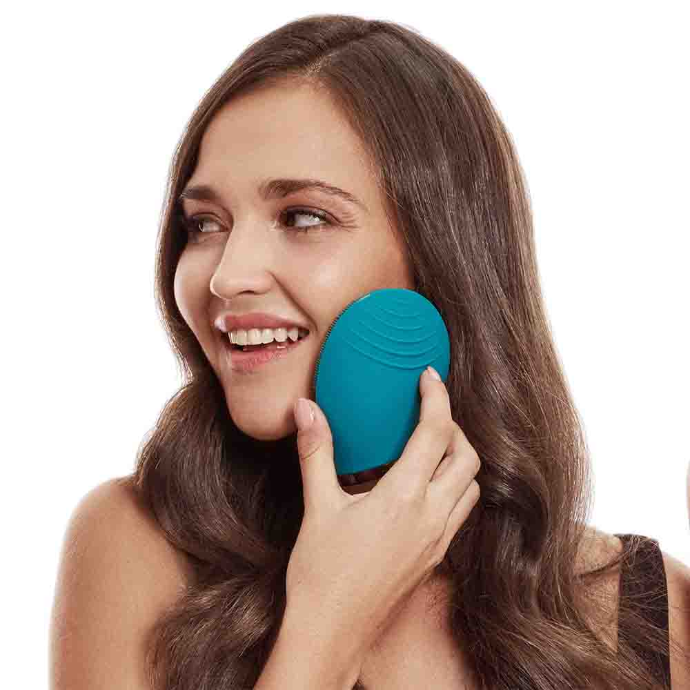 Mujer usando un dispositivo de limpieza facial turquesa en su mejilla, sonriendo.