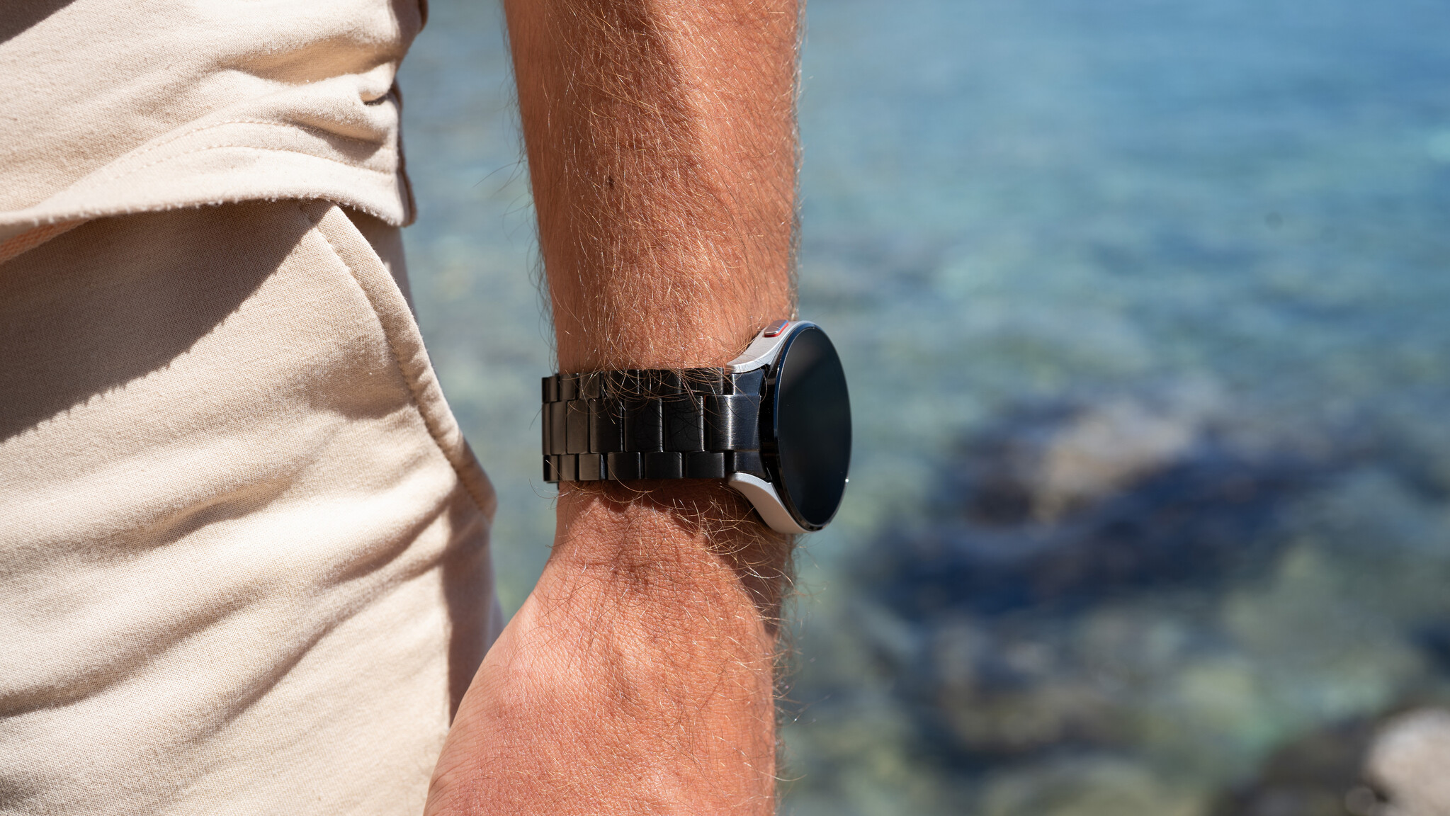 Handgelenk mit Uhr, schwarzem Armband und Wasser im Hintergrund.