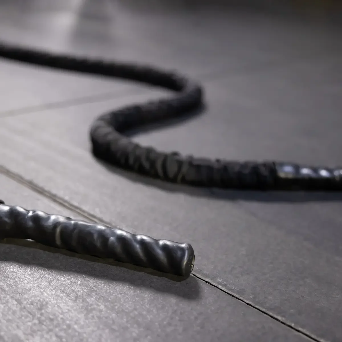 Black battle rope on a dark gray floor. The rope has a twisted design with a textured handle.