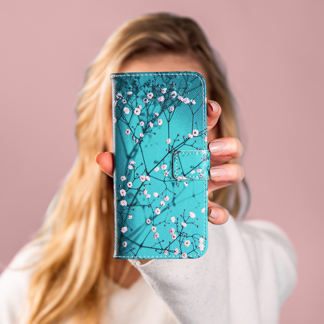Une femme tient un étui de téléphone turquoise avec des fleurs blanches. Cheveux blonds en arrière-plan.