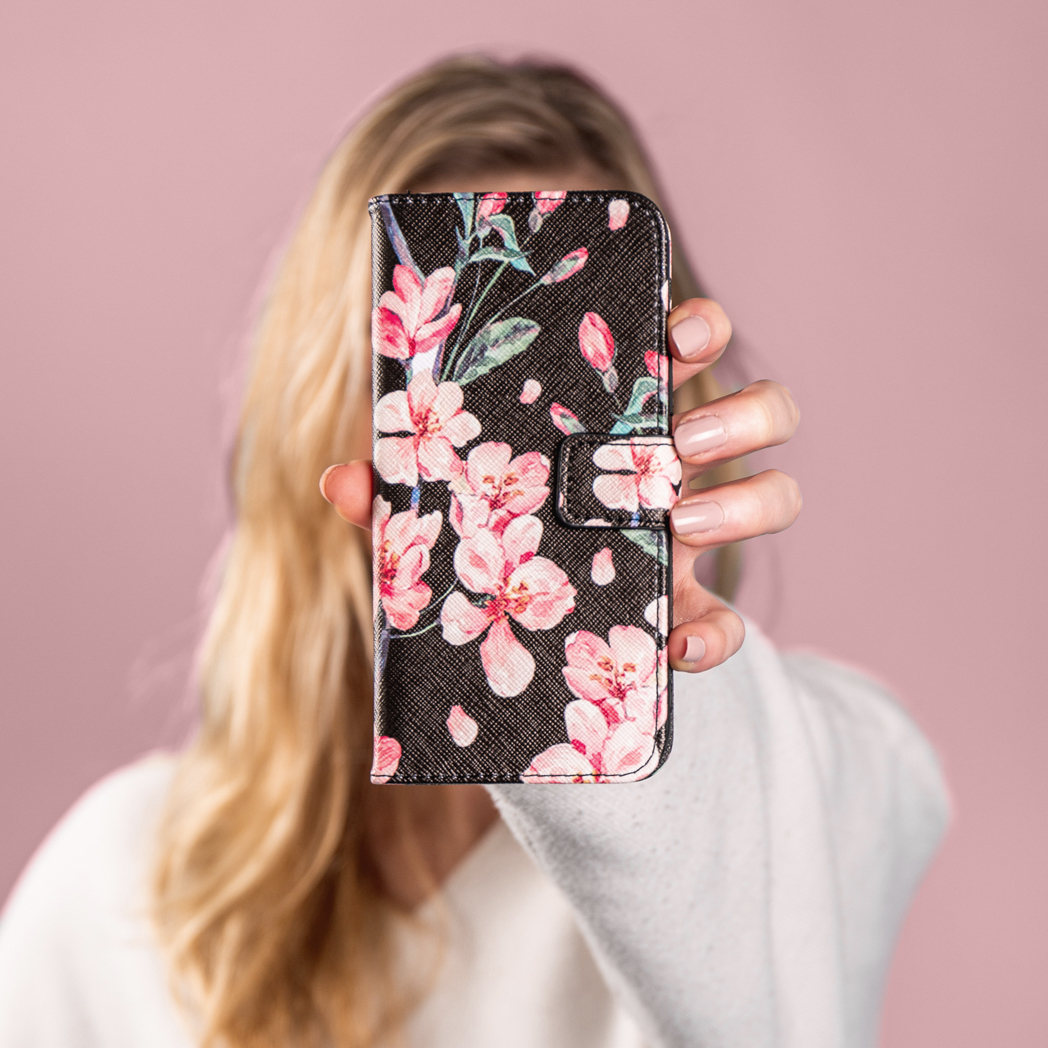 Een vrouw die een bloemen telefoonhoes voor haar gezicht houdt tegen een roze achtergrond.