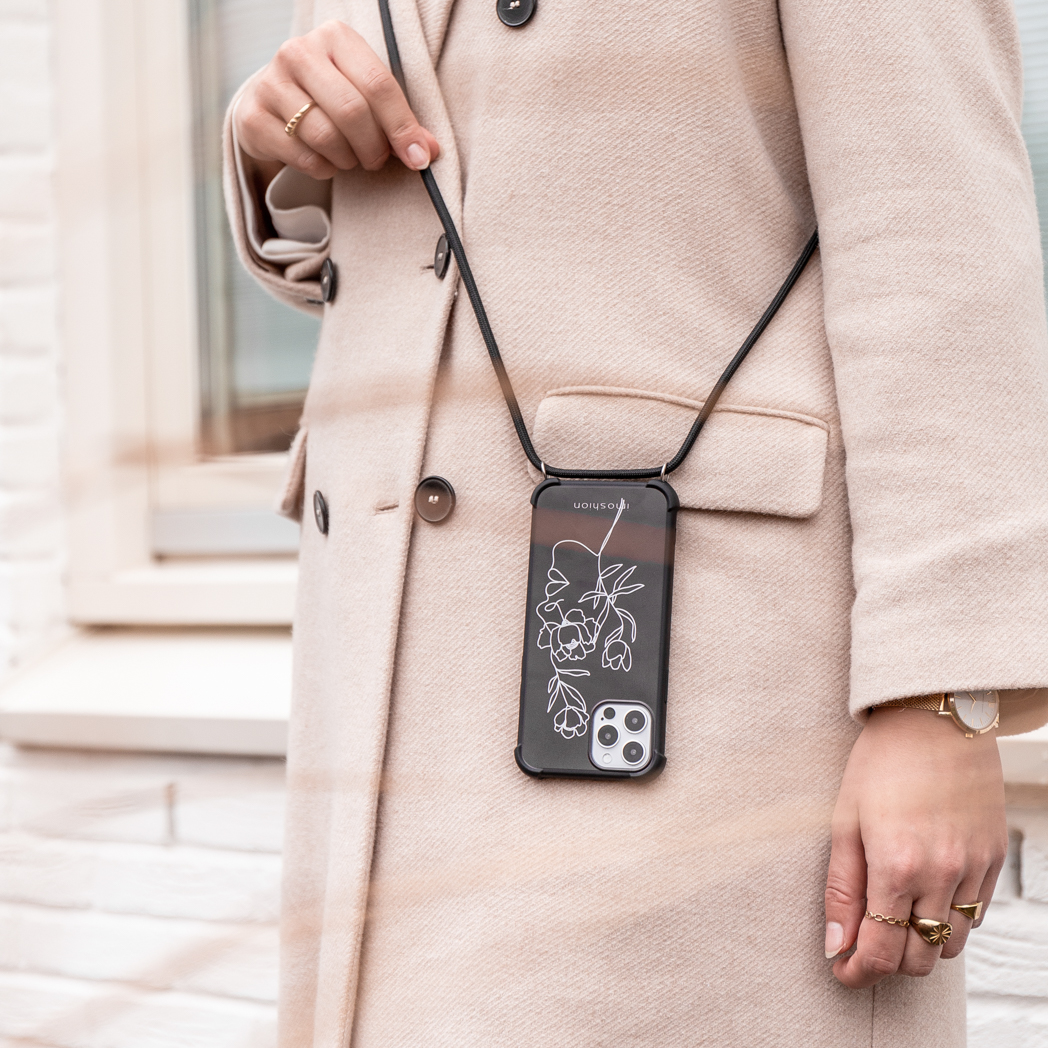 Een vrouw met een beige jas houdt een telefoon met een zwarte band vast, witte bakstenen muur.