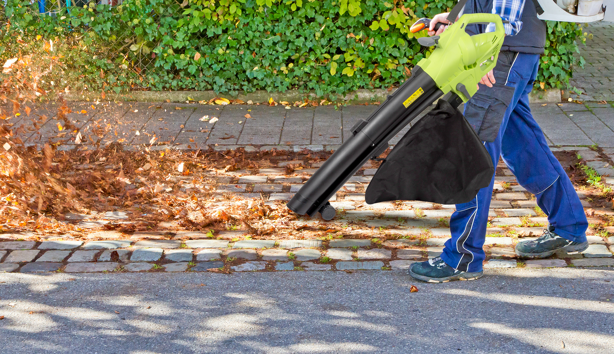 Een persoon in een blauwe broek en een zwarte jas gebruikt een bladblazer om bladeren op een stoep te blazen.