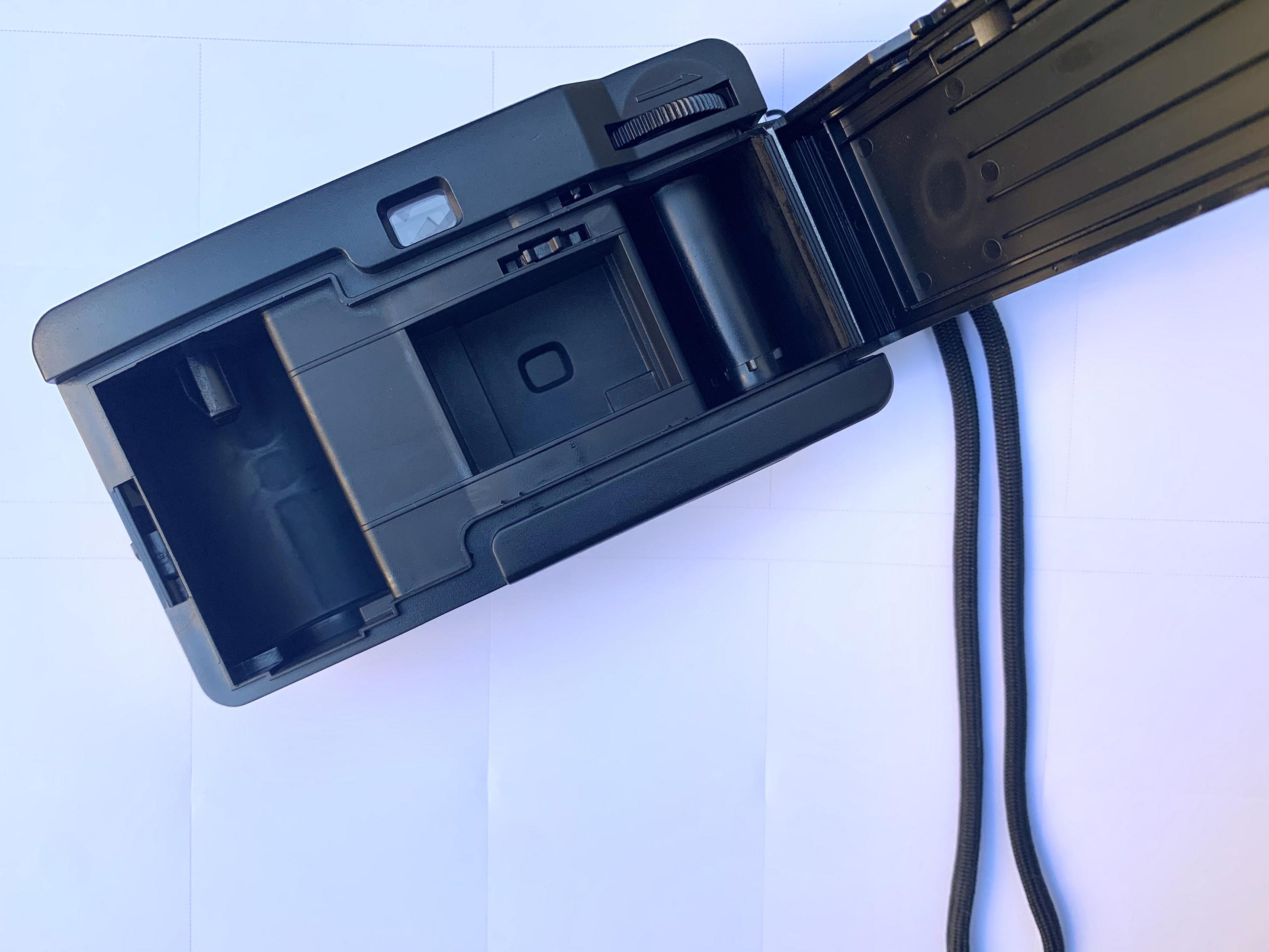 Otwarty czarny aparat z rolką filmu w środku i czarnym paskiem. Aparat na białej powierzchni.