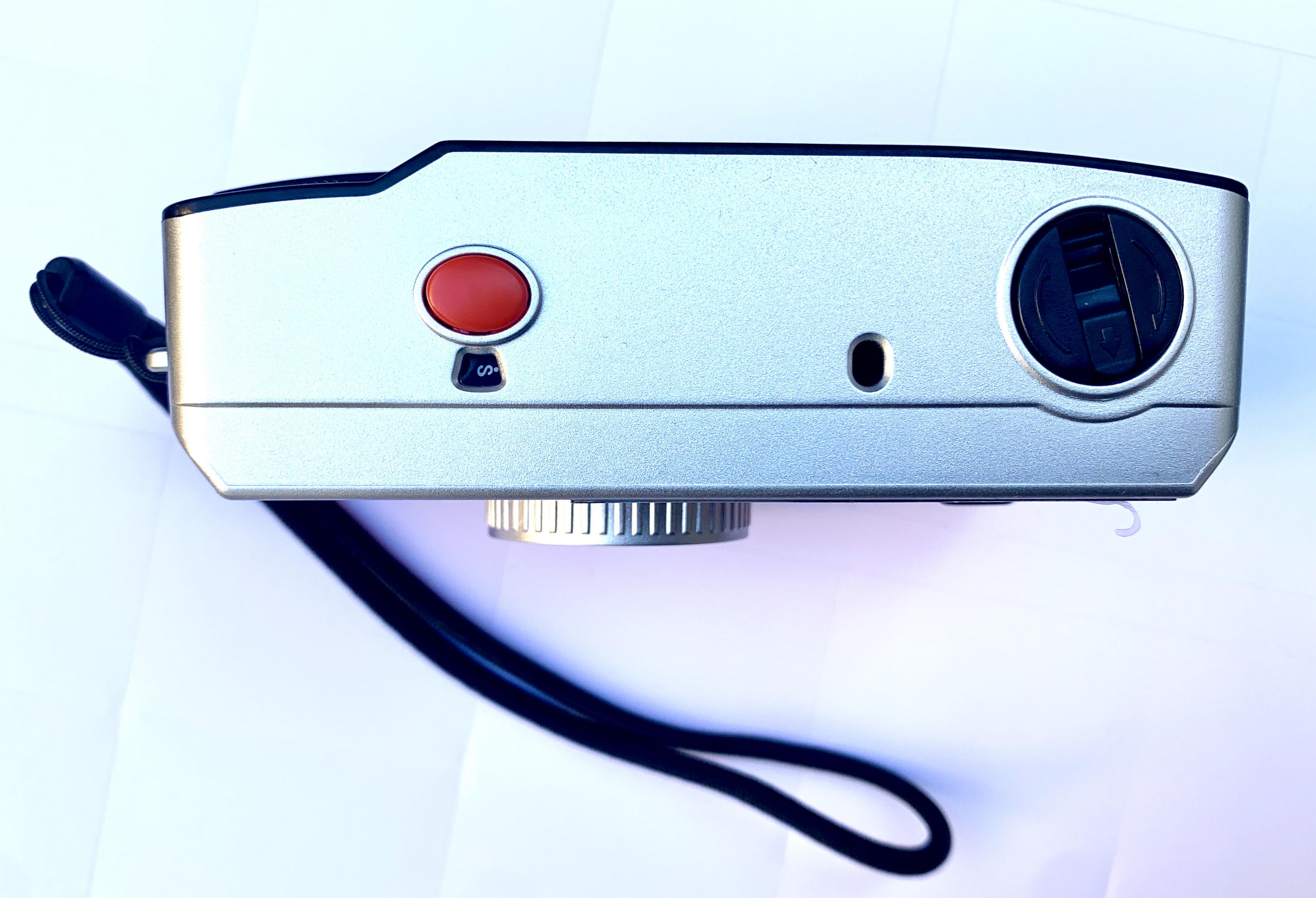 Srebrny aparat fotograficzny z góry, z czerwonym przyciskiem, paskiem i czarnymi detalami.