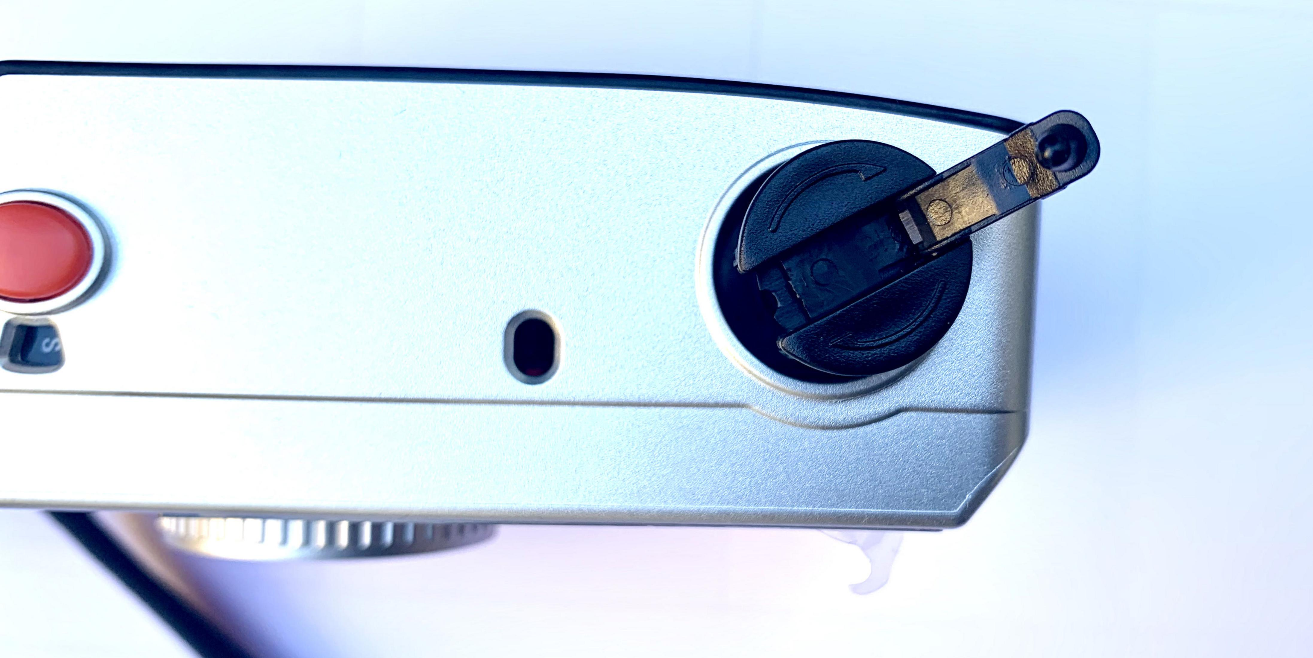 Zbliżenie srebrnego aparatu z czarnym pokrętłem u góry i czerwonym przyciskiem po lewej stronie.