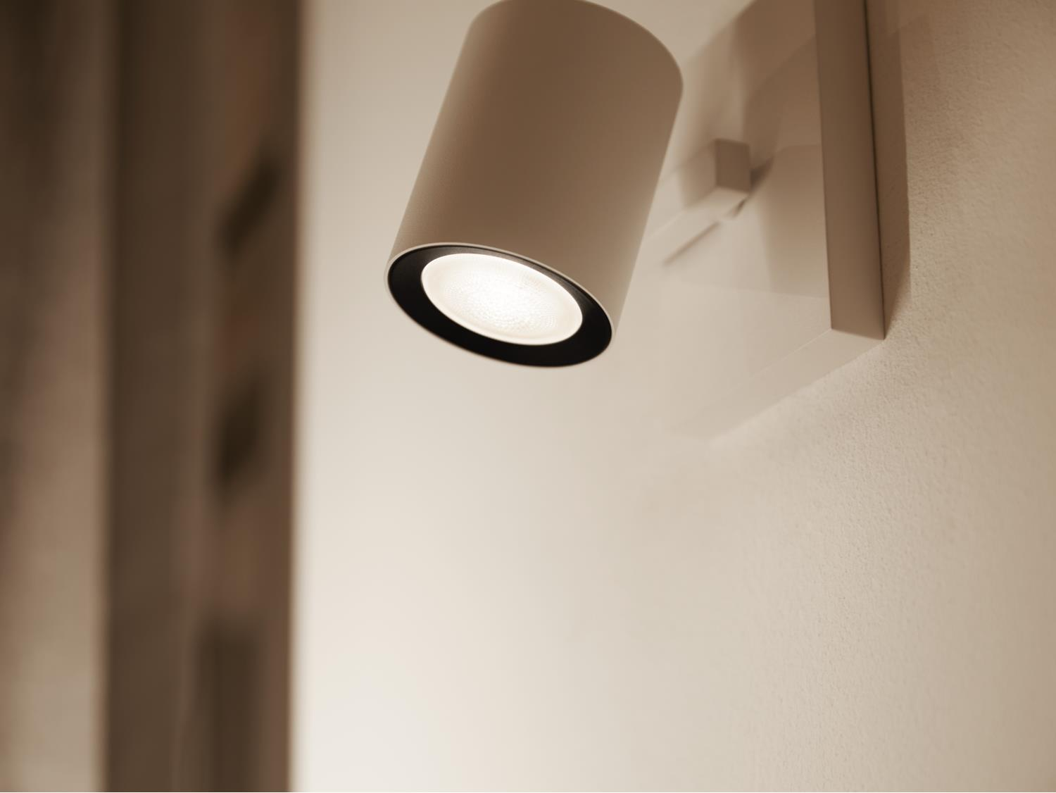 Weiße moderne Wandleuchte mit zylindrischem Kopf. Das Licht ist an und wirft einen Schein. Braune Wand.