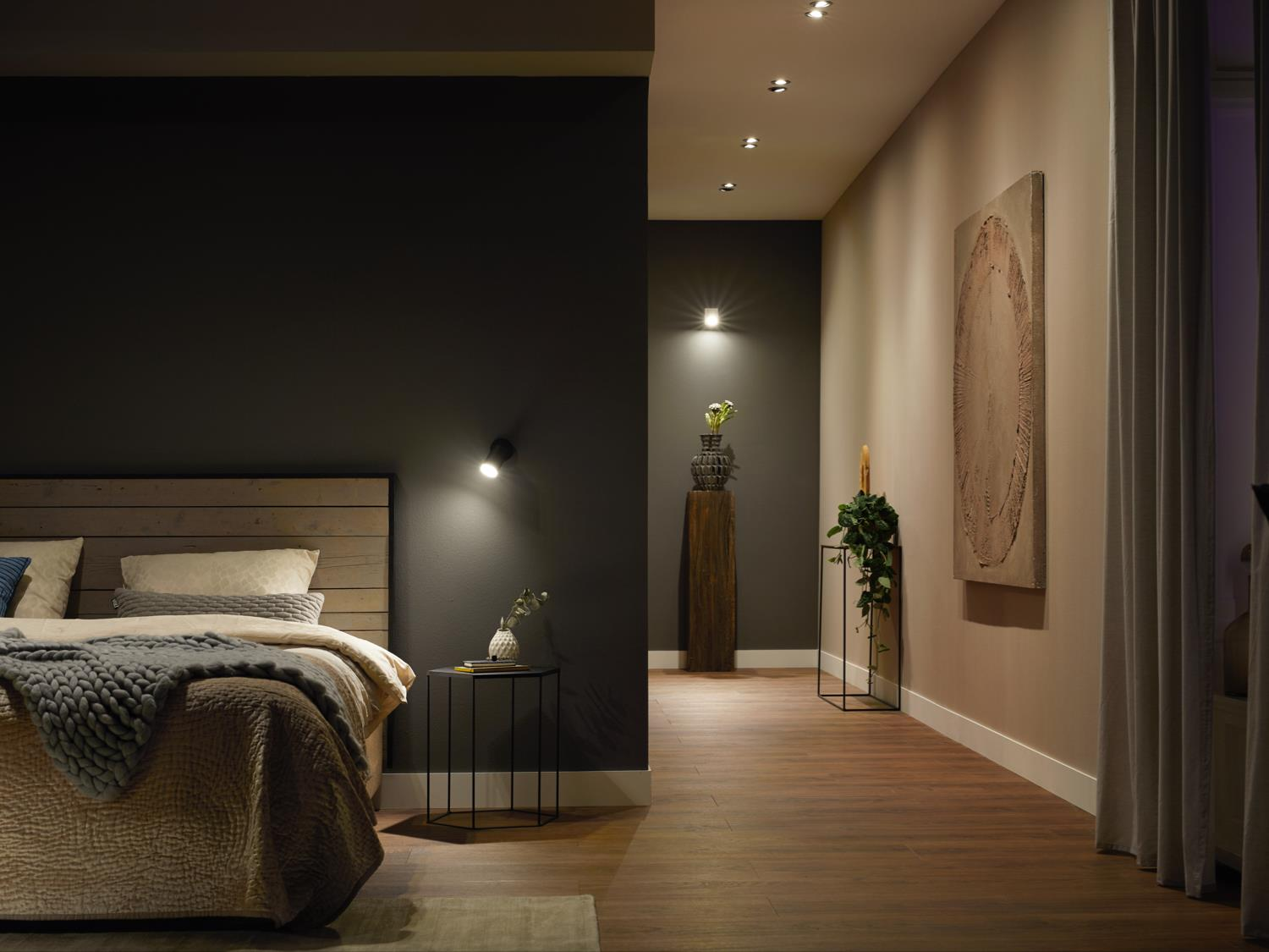 Ein Schlafzimmerinterieur mit Strahlern, einem Bett und Pflanzen, mit dunkler Wand und Holzboden.