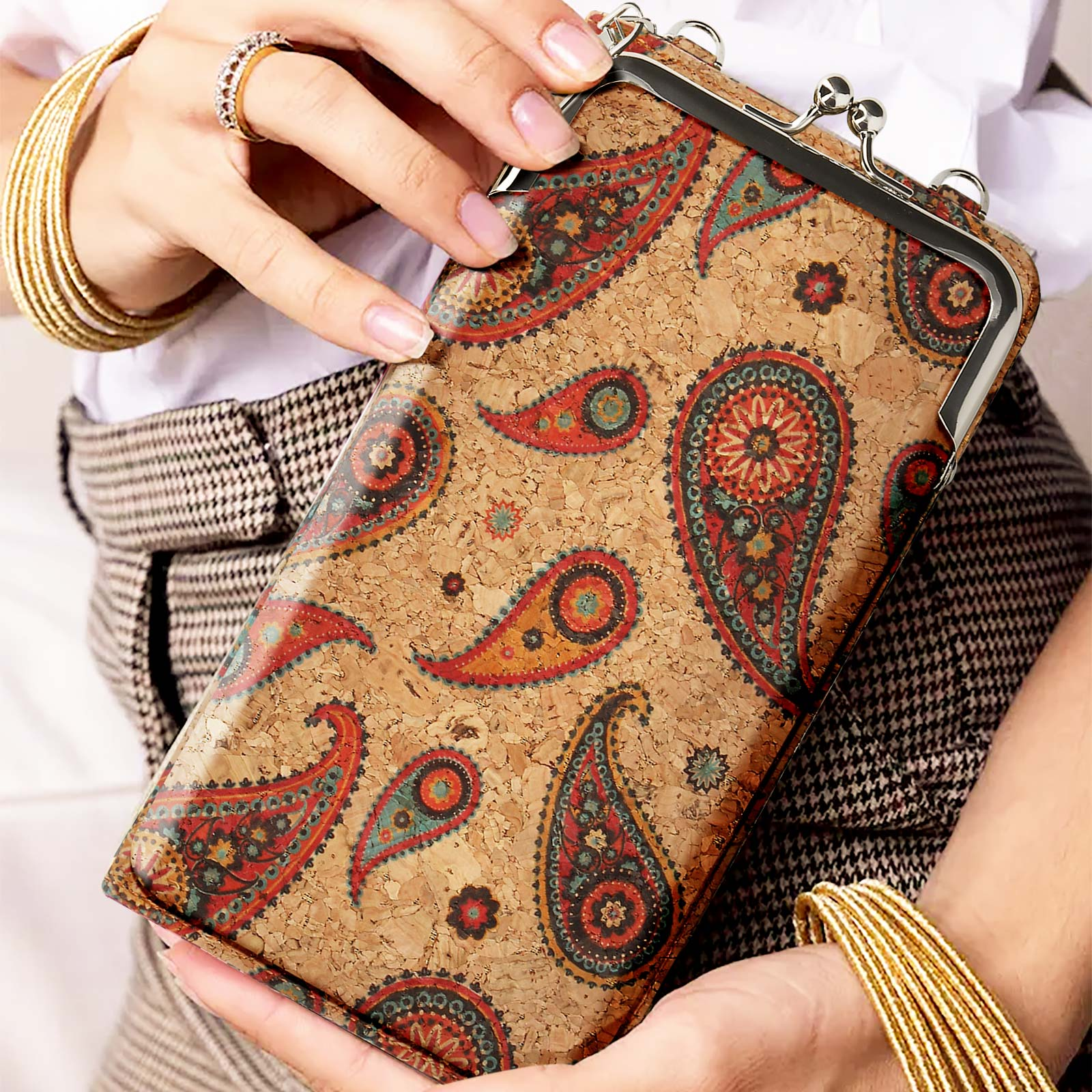 Frau hält eine braune Korkbrieftasche mit Paisley-Design. Sie trägt goldene Armbänder und einen Ring.