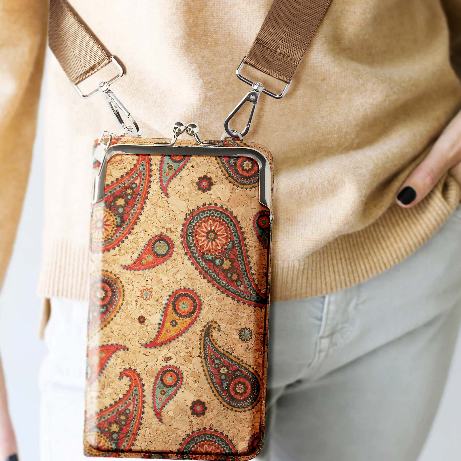 Eine braune Paisley-Geldbörse mit Riemen hängt herunter. Die Person trägt einen beigefarbenen Pullover und helle Hosen.