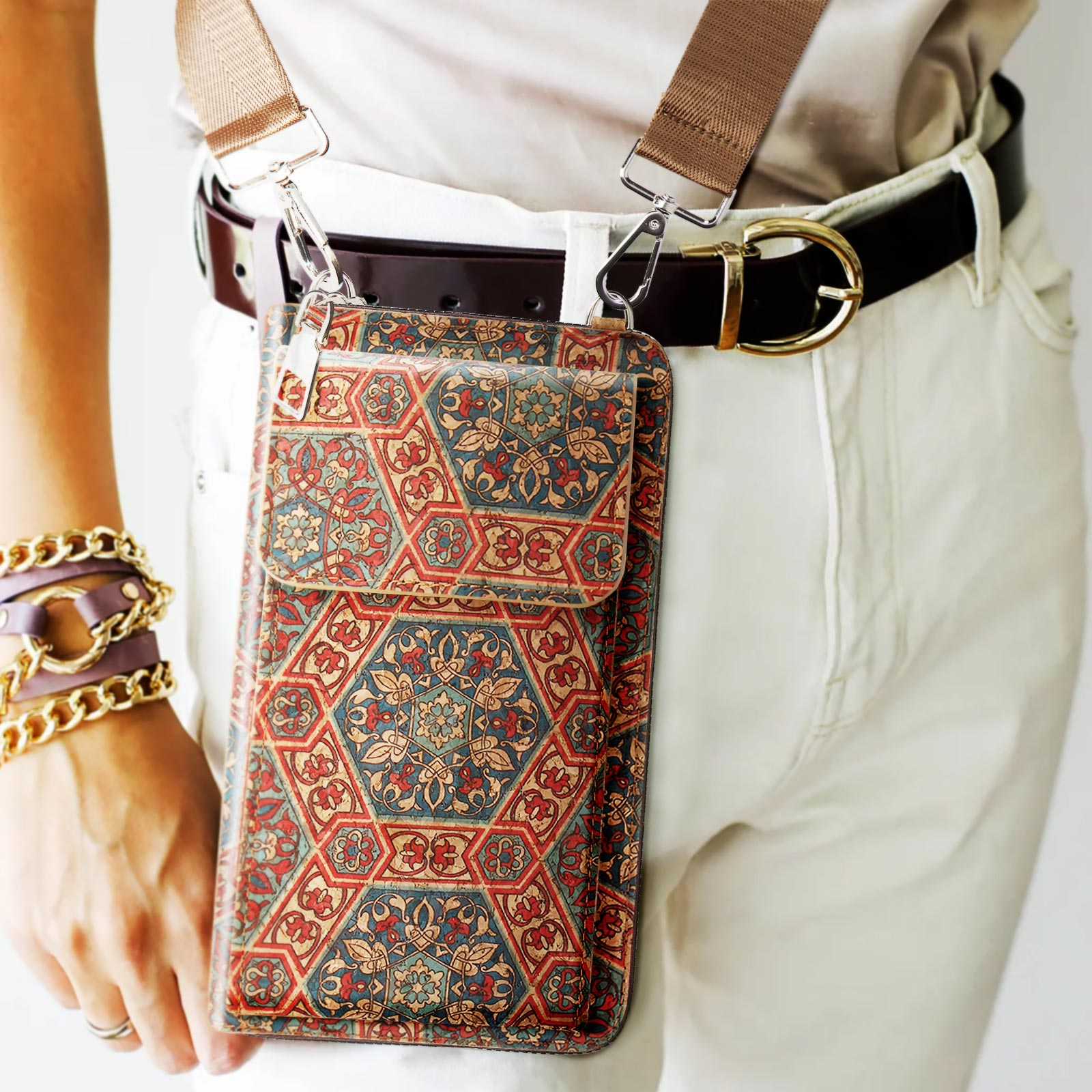 Person trägt eine Crossbody-Telefontasche mit buntem Blumendesign. Weiße Hose und brauner Gürtel.