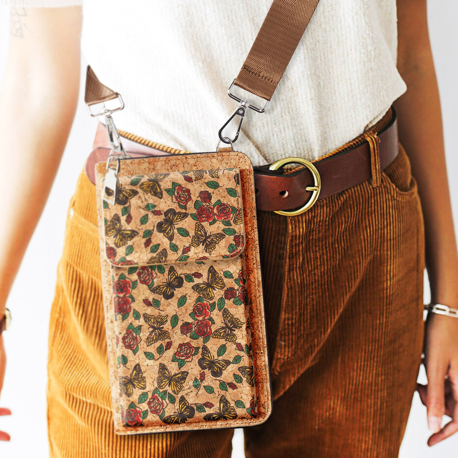 Person trägt eine Crossbody-Handyhalterung mit Schmetterlings- und Rosenmuster. Brauner Gürtel und Cordhose.