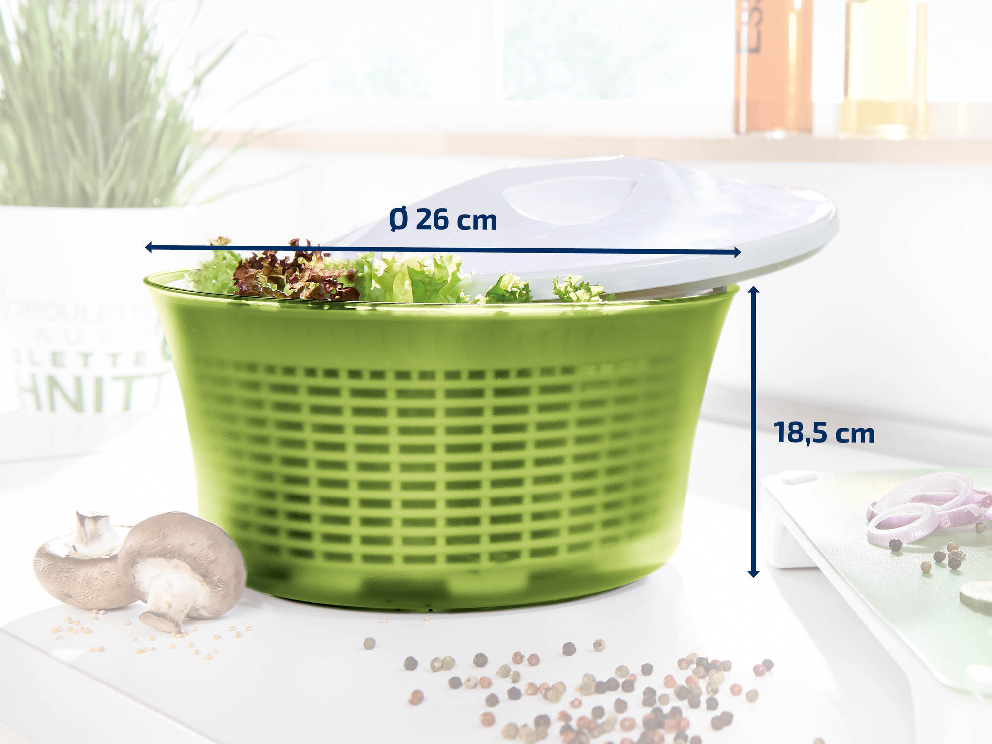 Grüner Salatschleuder mit Salat darin. Abmessungen sind beschriftet. Pilze und Pfefferkörner sind in der Szene.