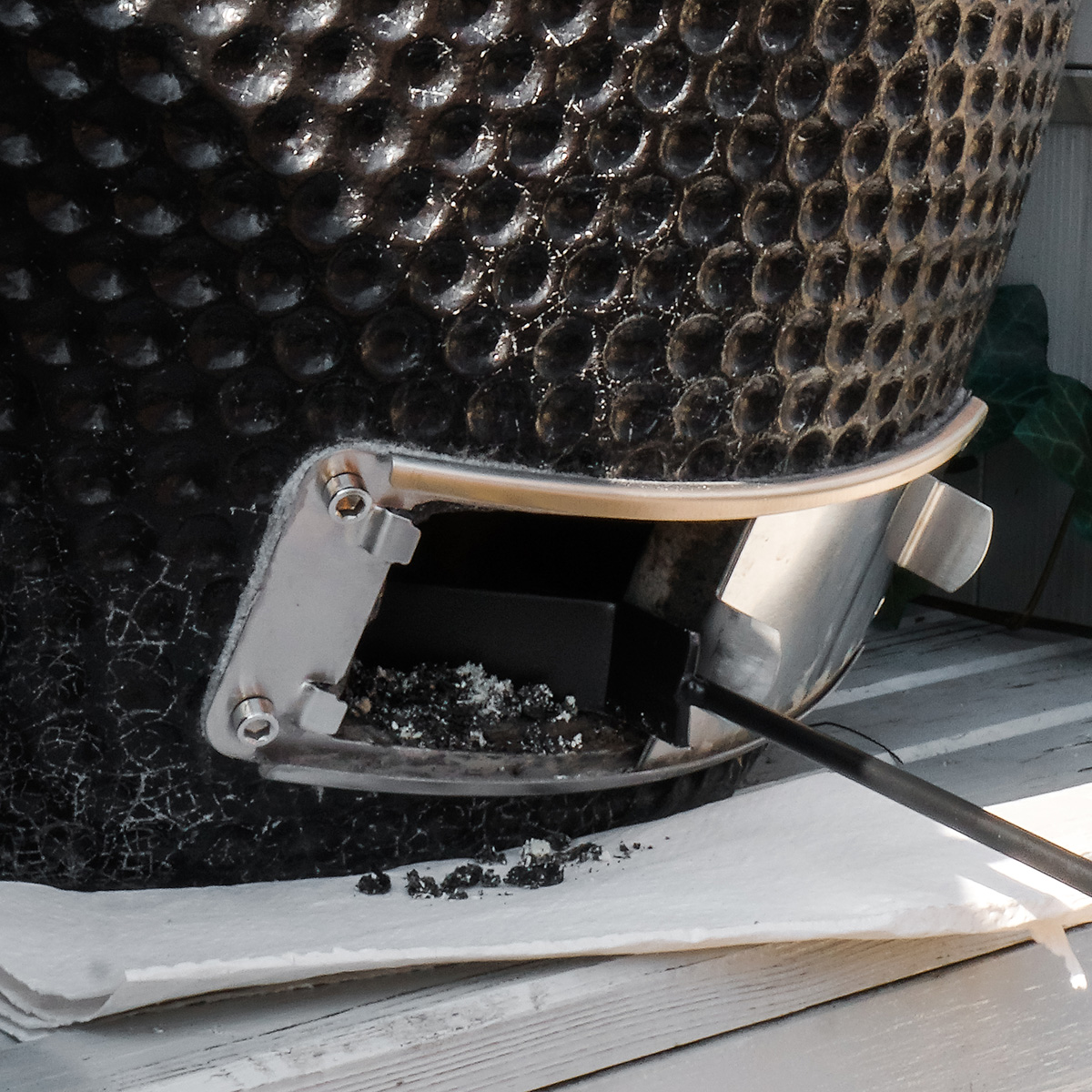 A black grill with ash and a tool inside, on a white surface with a wooden background.