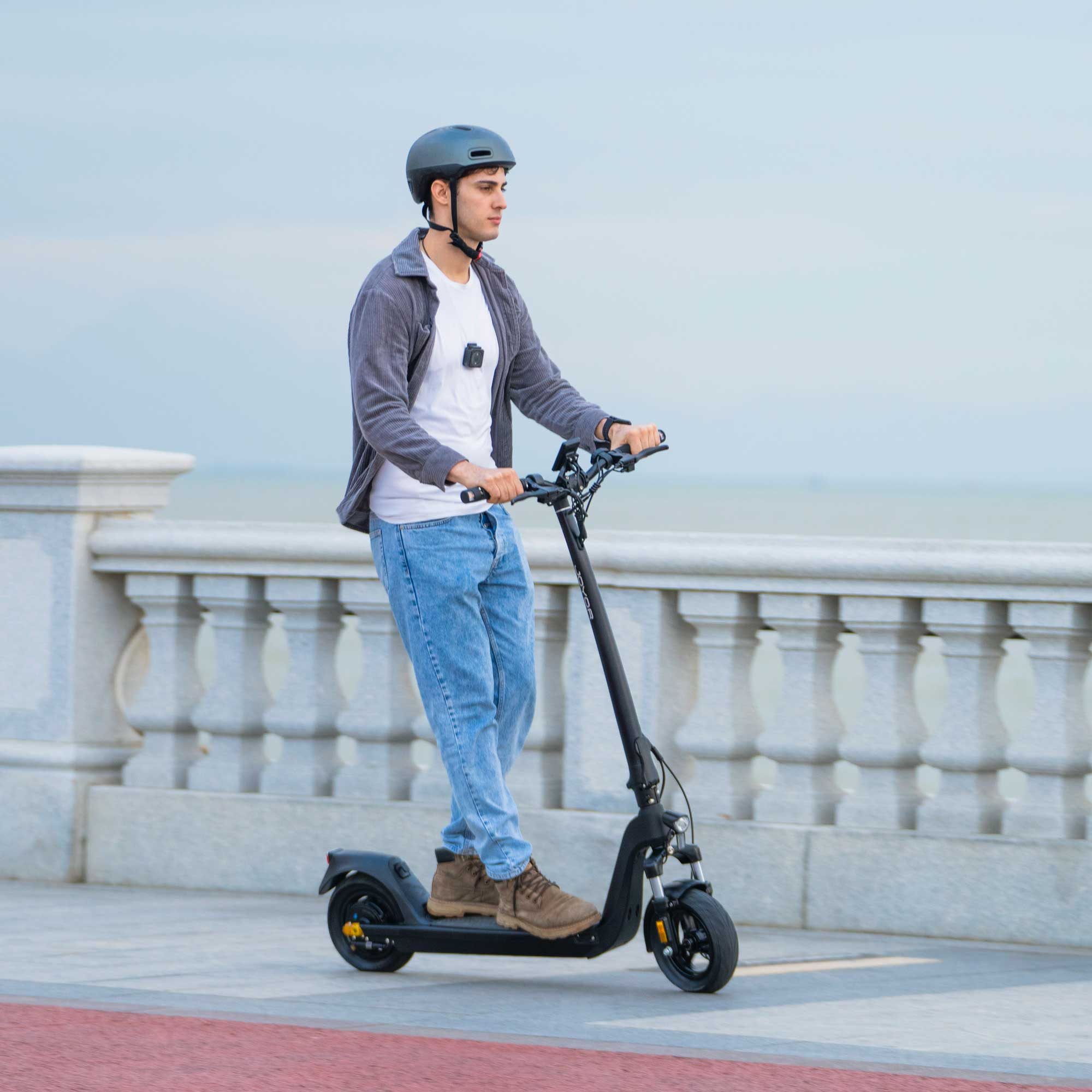 Ein Mann fährt mit einem schwarzen Elektroroller auf einem gepflasterten Weg. Er trägt einen Helm und Jeans.