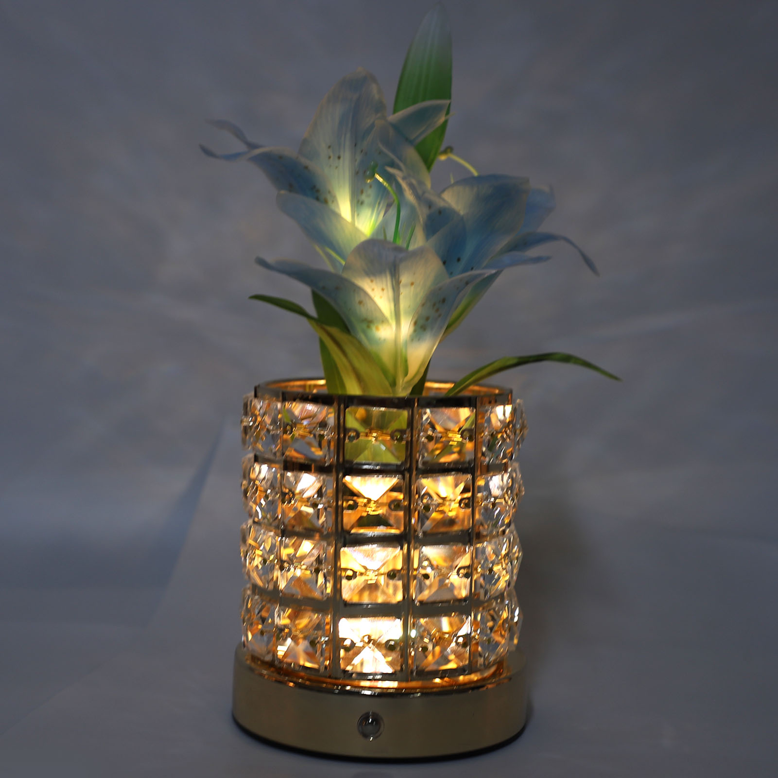 Goldene Kristallvase mit hellblauen Blütenstielen und -blüten. Lichter sind an.