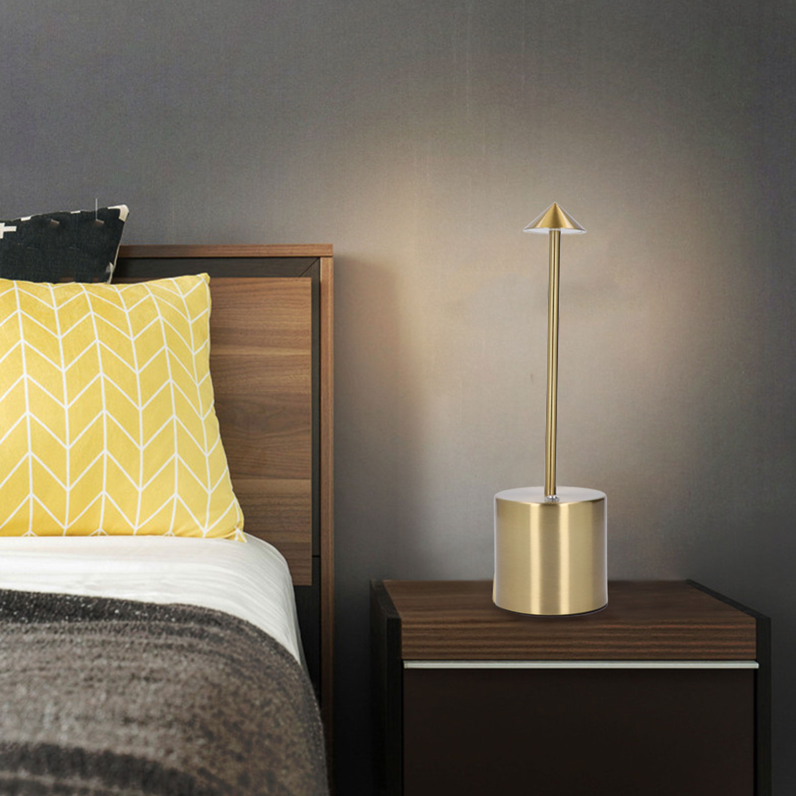 Eine goldene Lampe auf einem Nachttisch, mit einem gelben Kissen und einem Bett im Hintergrund.
