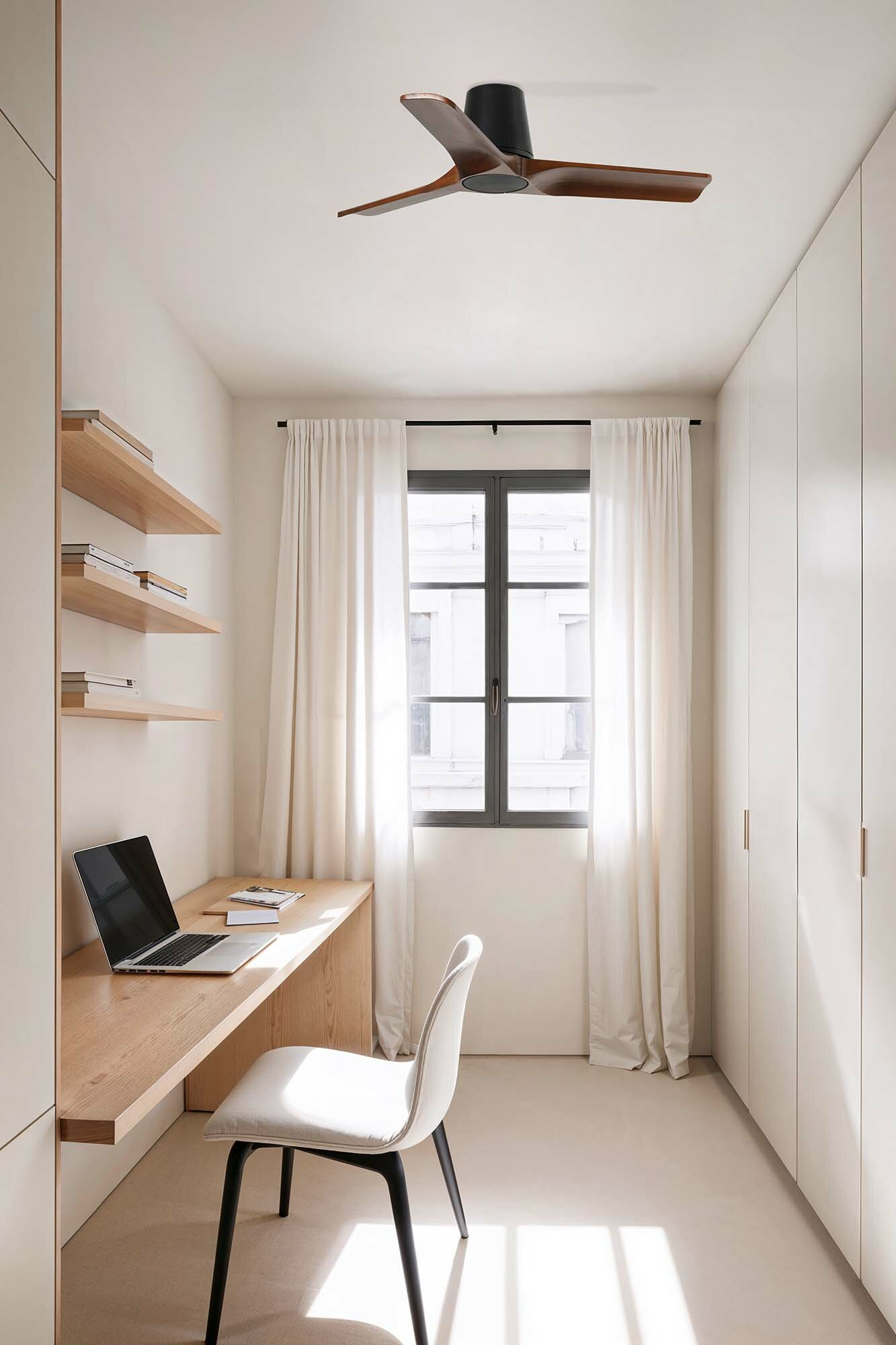 Innenraum eines Arbeitszimmers mit Holztisch, Regalen, Laptop und Fenster mit Vorhängen. Weiße Wände.