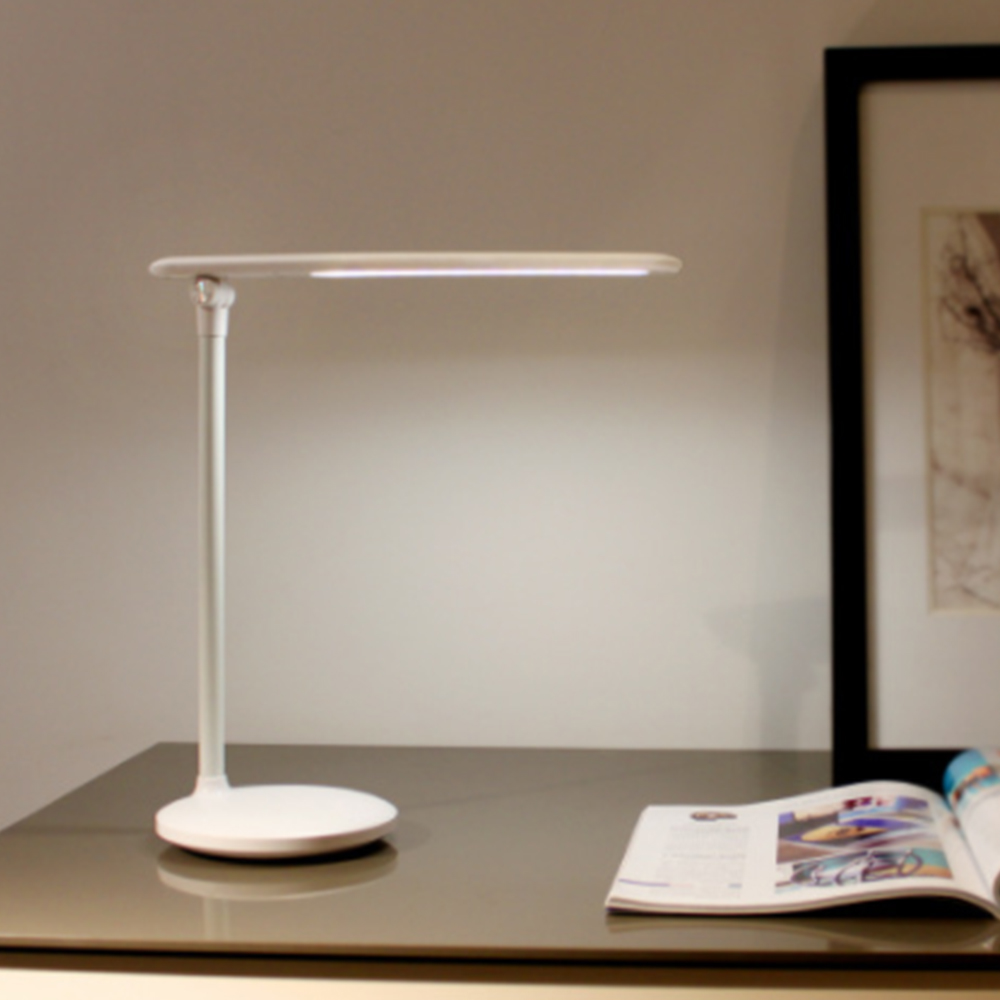 Eine weiße Schreibtischlampe auf einem Tisch. Ein Magazin ist neben der Lampe geöffnet. Weiße Wand im Hintergrund.