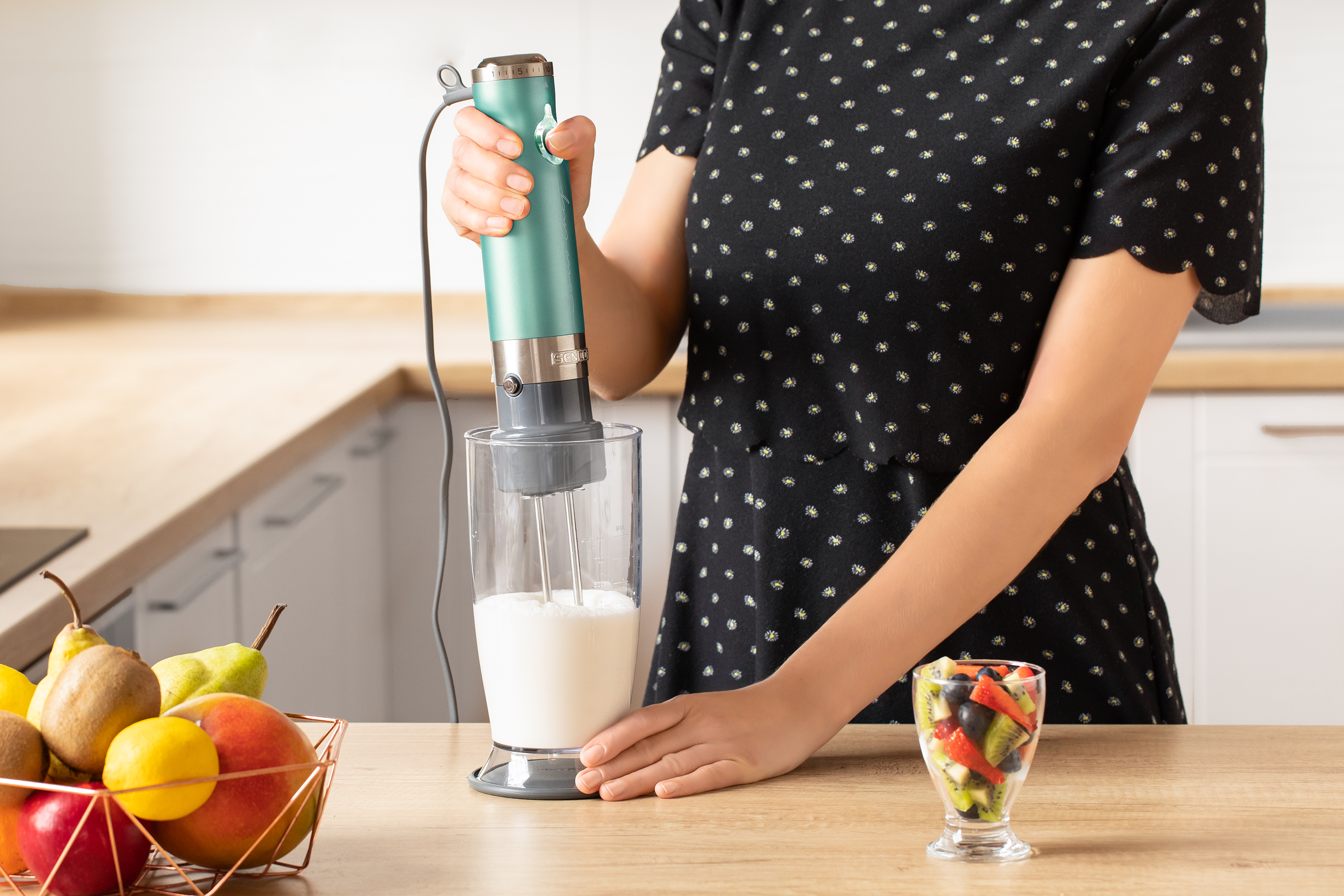 Donna usa un frullatore a immersione verde acqua per mescolare liquido in un bicchiere. Frutta sul tavolo.