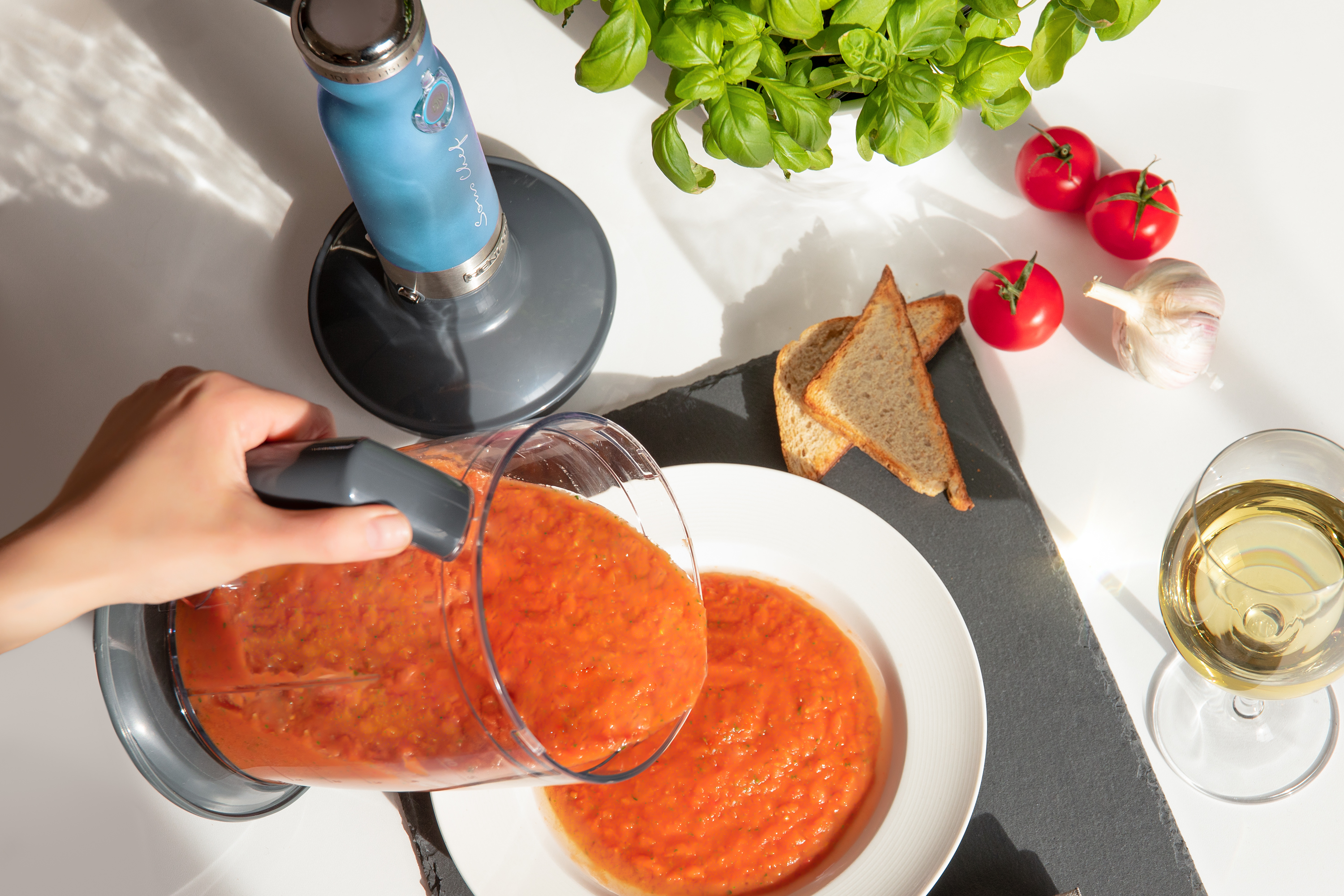 Una mano versa zuppa arancione da un frullatore in una ciotola bianca. Ci sono pomodori e basilico vicino.