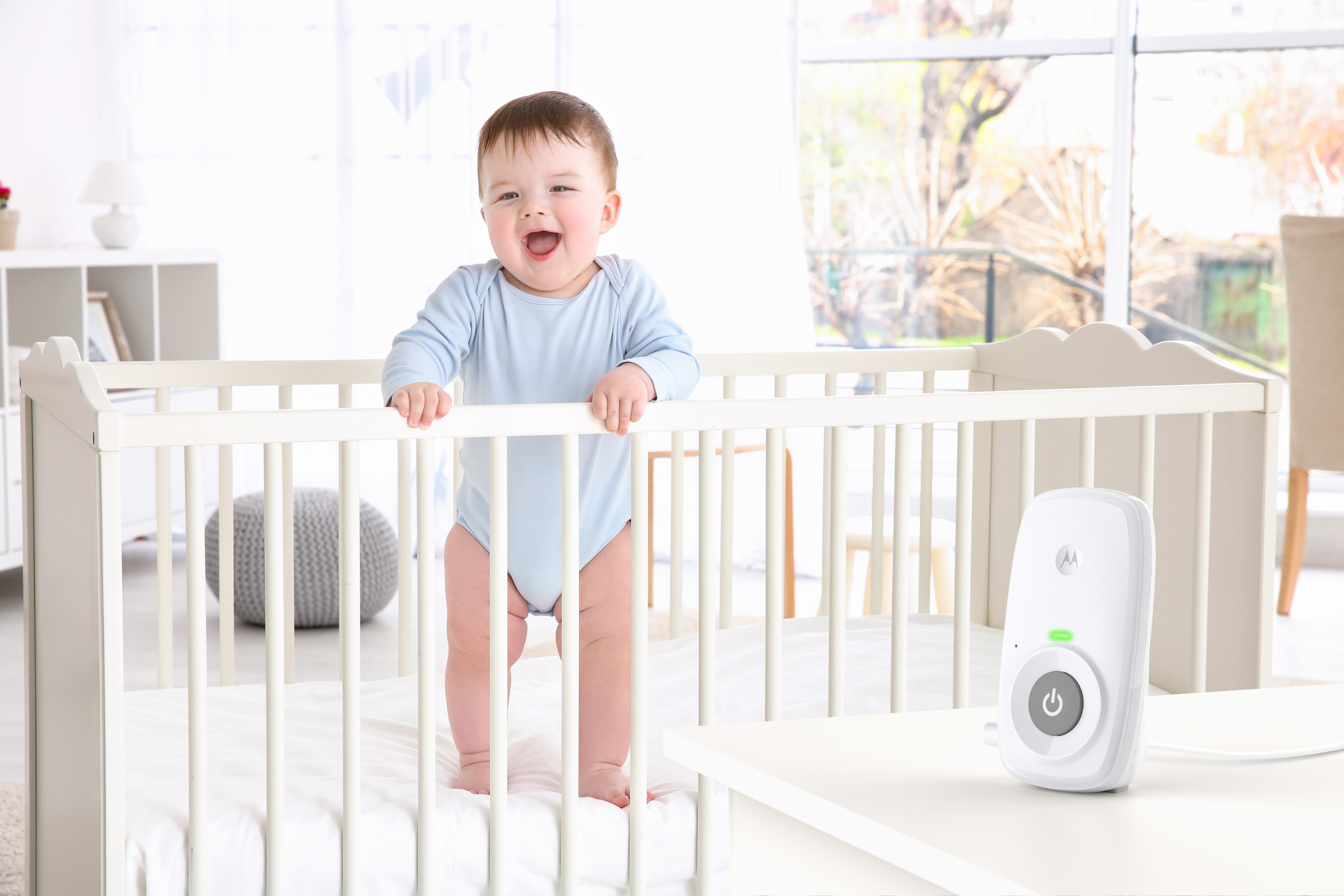 Dziecko w łóżeczku uśmiecha się, z nianią na stole. Pokój ma okno.