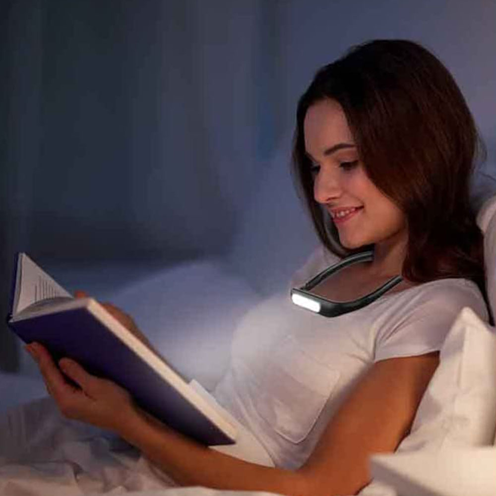 Eine Frau liest ein Buch mit einer Nackenleuchte. Sie liegt im Bett mit weißen Kissen.