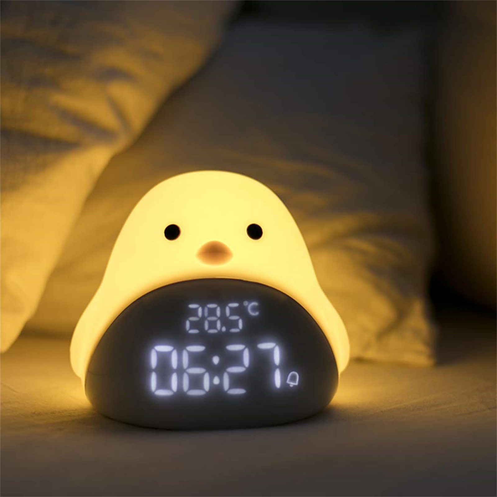 Ein gelber pinguinförmiger Wecker, der Zeit und Temperatur anzeigt. Er liegt auf einem Bett.
