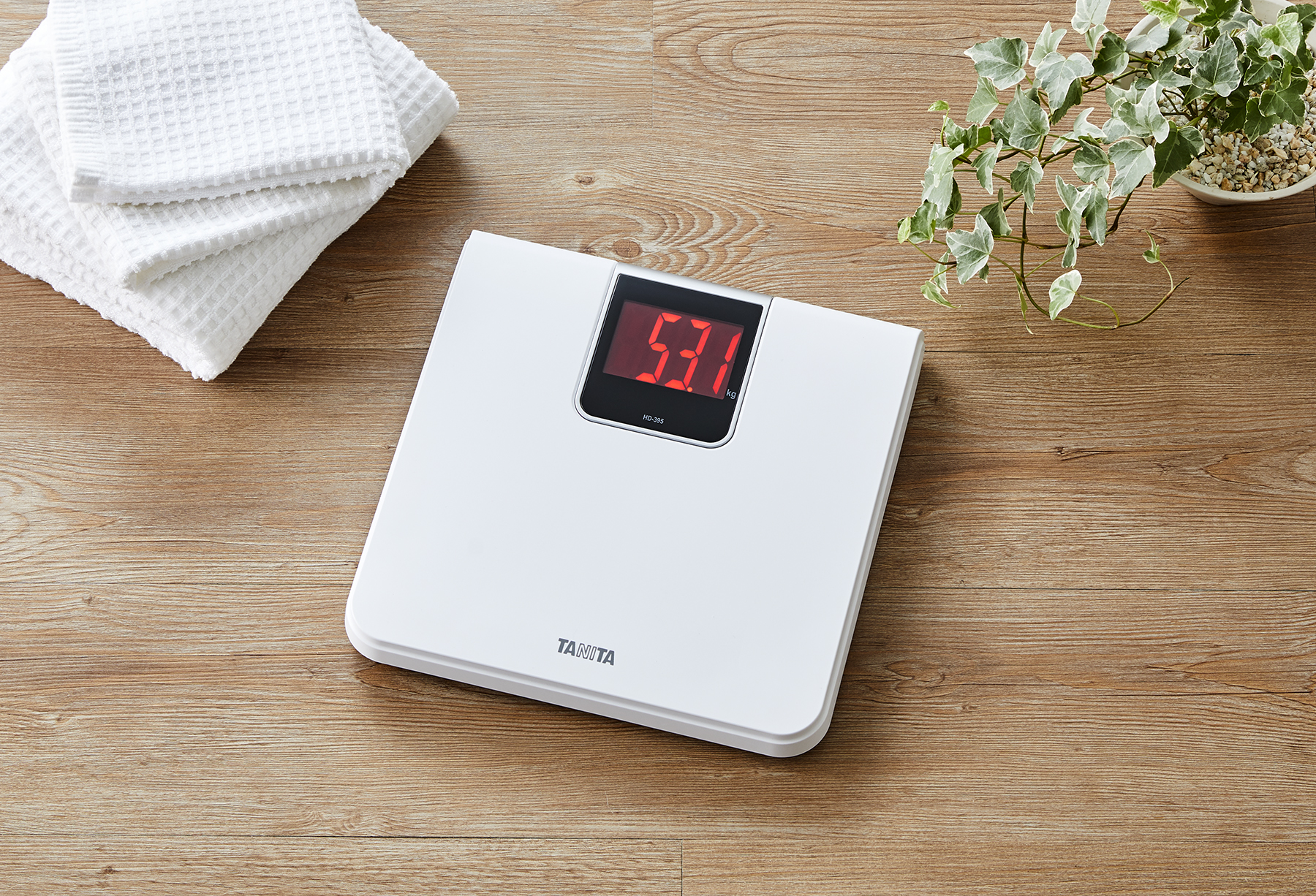 Een witte digitale weegschaal met 53,1 kg, geplaatst op een houten tafel met handdoeken en een plant.