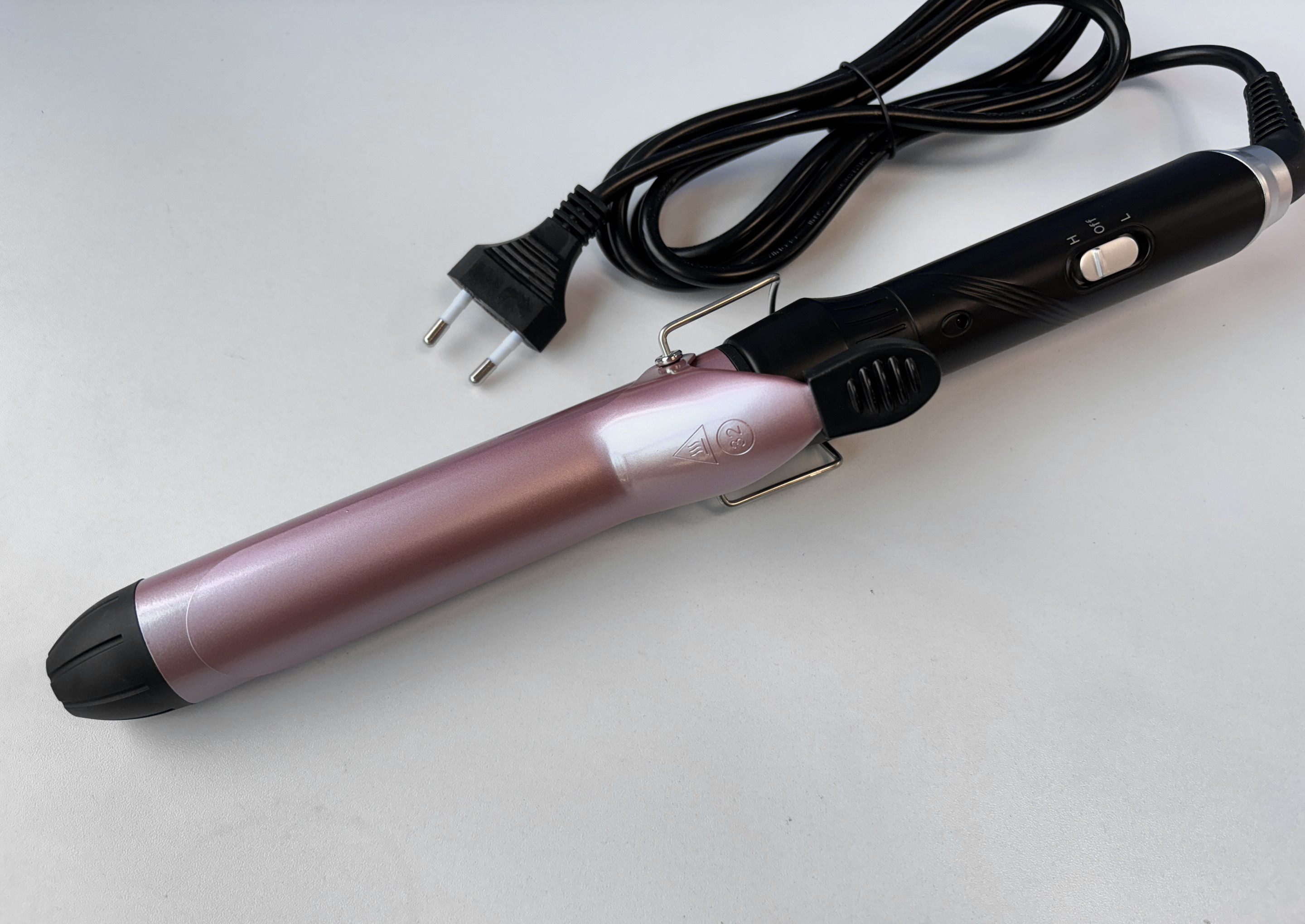 Un ferro arricciacapelli con un fusto rosa, manico nero e cavo di alimentazione. Sfondo bianco e una spina.
