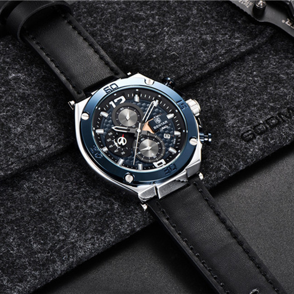 Eine Uhr mit blauem und silbernem Zifferblatt und schwarzem Lederarmband. Die Uhr liegt auf einer grauen Oberfläche. Der Hintergrund ist dunkel.