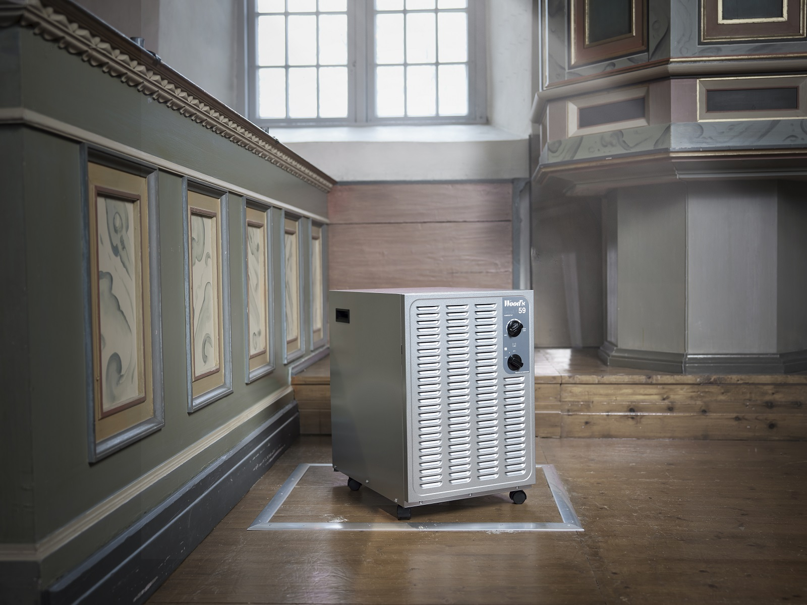 Ein silberner Luftreiniger auf Rädern in einer Kirche. Zwei Bedienknöpfe. Holzwände und -boden, Fenster.