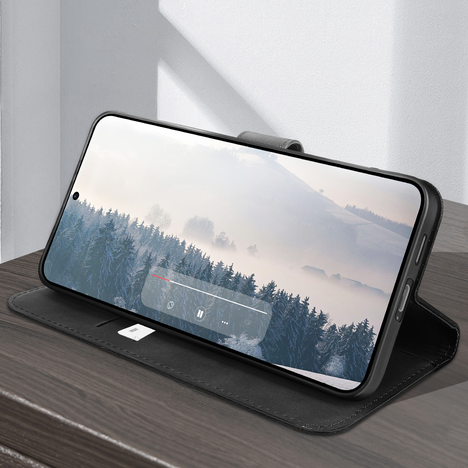 Un telefono in una custodia nera su un tavolo di legno. Lo schermo del telefono mostra una scena di foresta invernale con un pulsante di riproduzione.