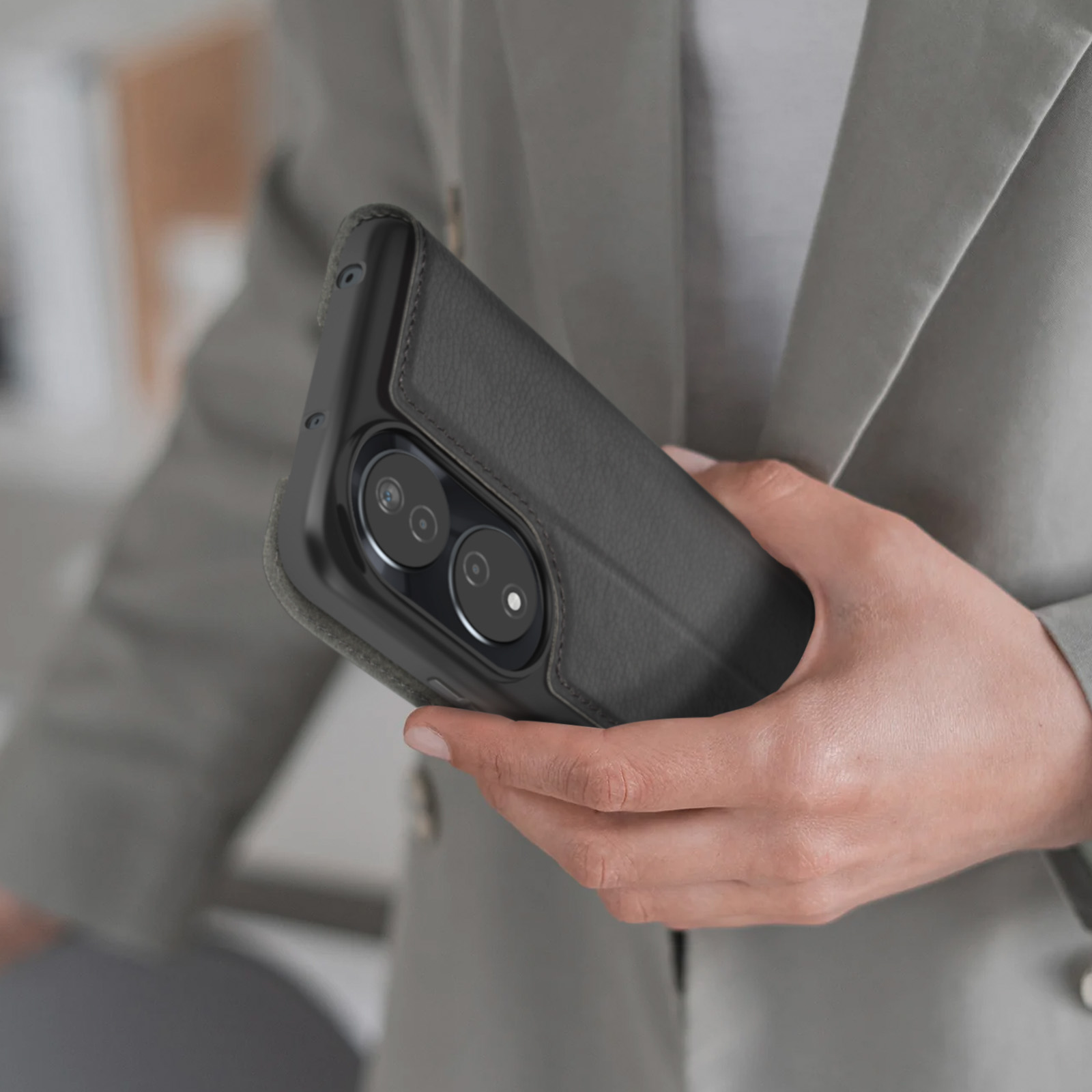 Hand hält geschlossene dunkelgraue Handyhülle mit Dual-Kamera-Ausschnitt. Person trägt grauen Blazer.