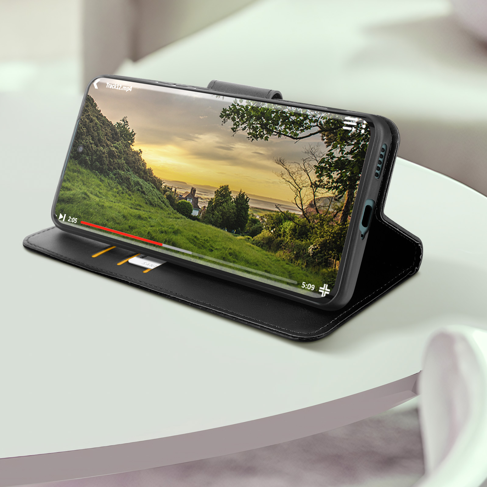Telefon auf schwarzer Hülle, spielt Video, zeigt Landschaft. Weißer Tisch.