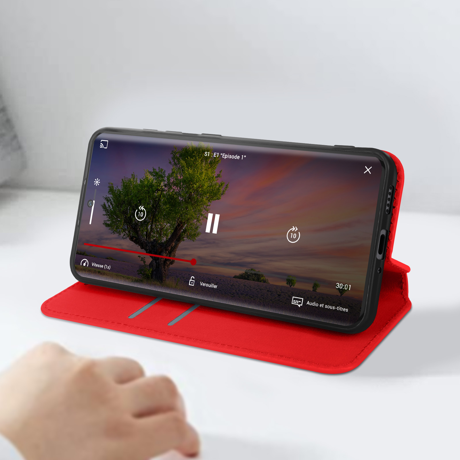 Telefono in custodia rossa, che riproduce video, superficie bianca, mano a sinistra.