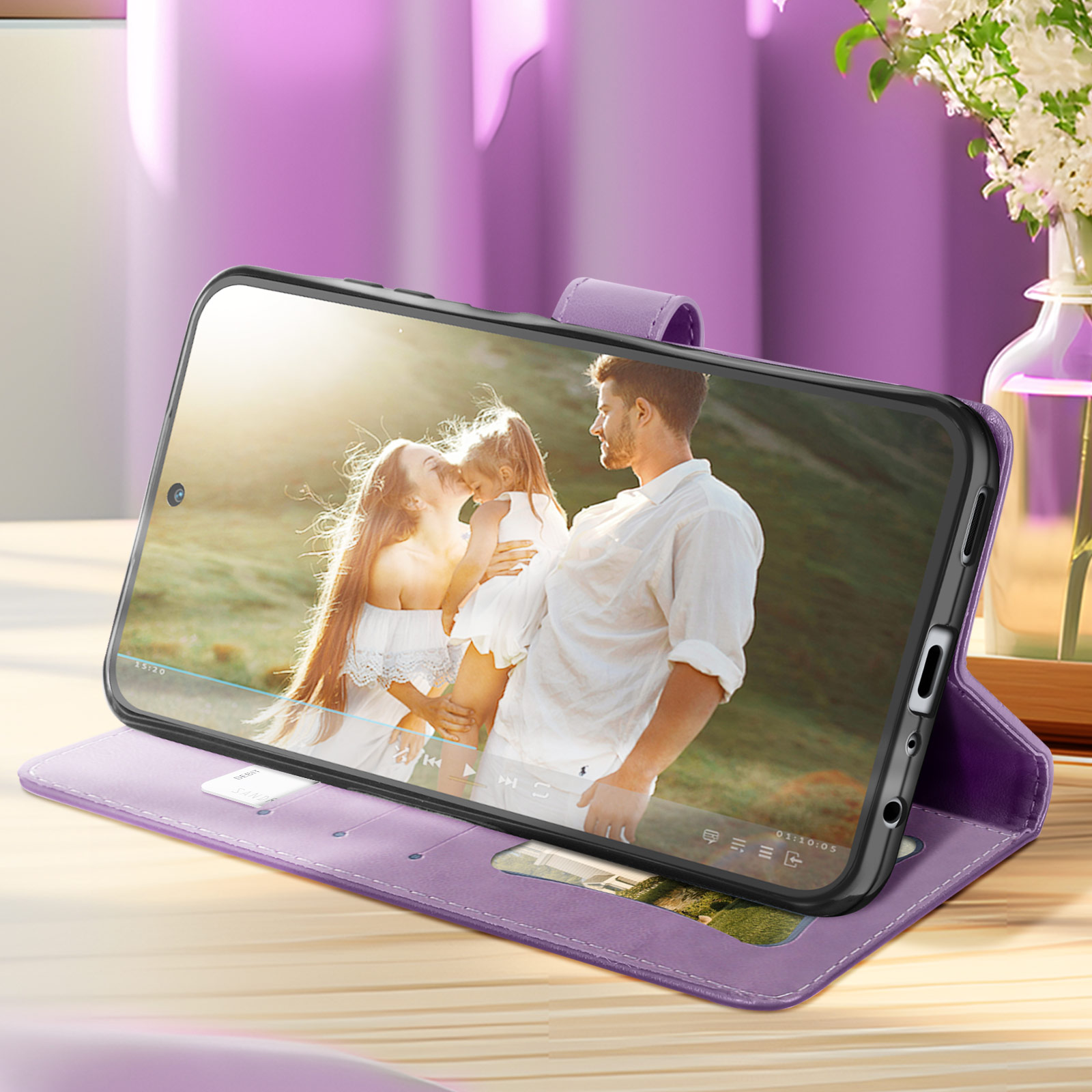 Telefono in custodia viola, mostra video di famiglia. Vaso e fiori sullo sfondo.