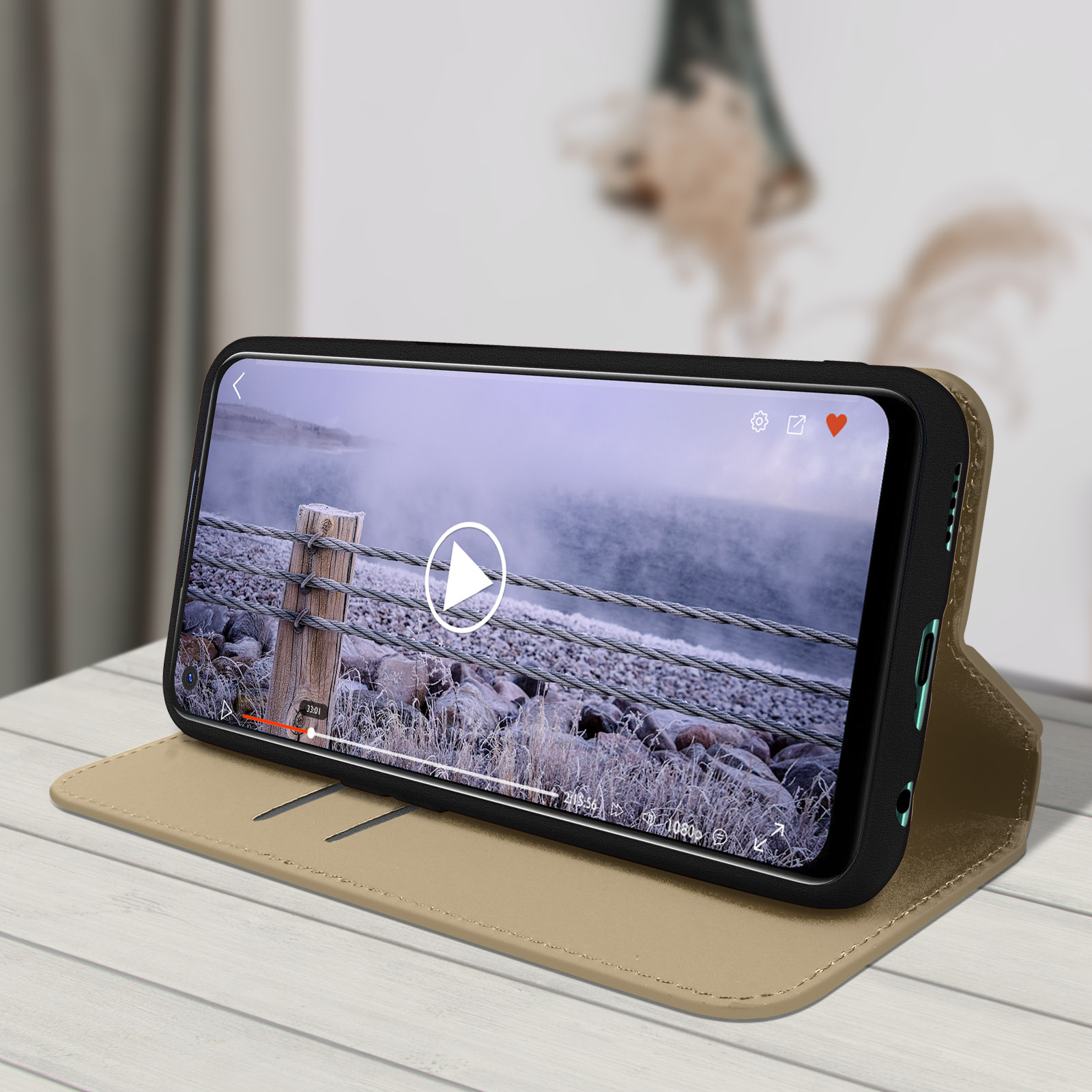 Telefono in custodia dorata, mostra un video, su superficie in legno. Sfondo: paesaggio sfocato.
