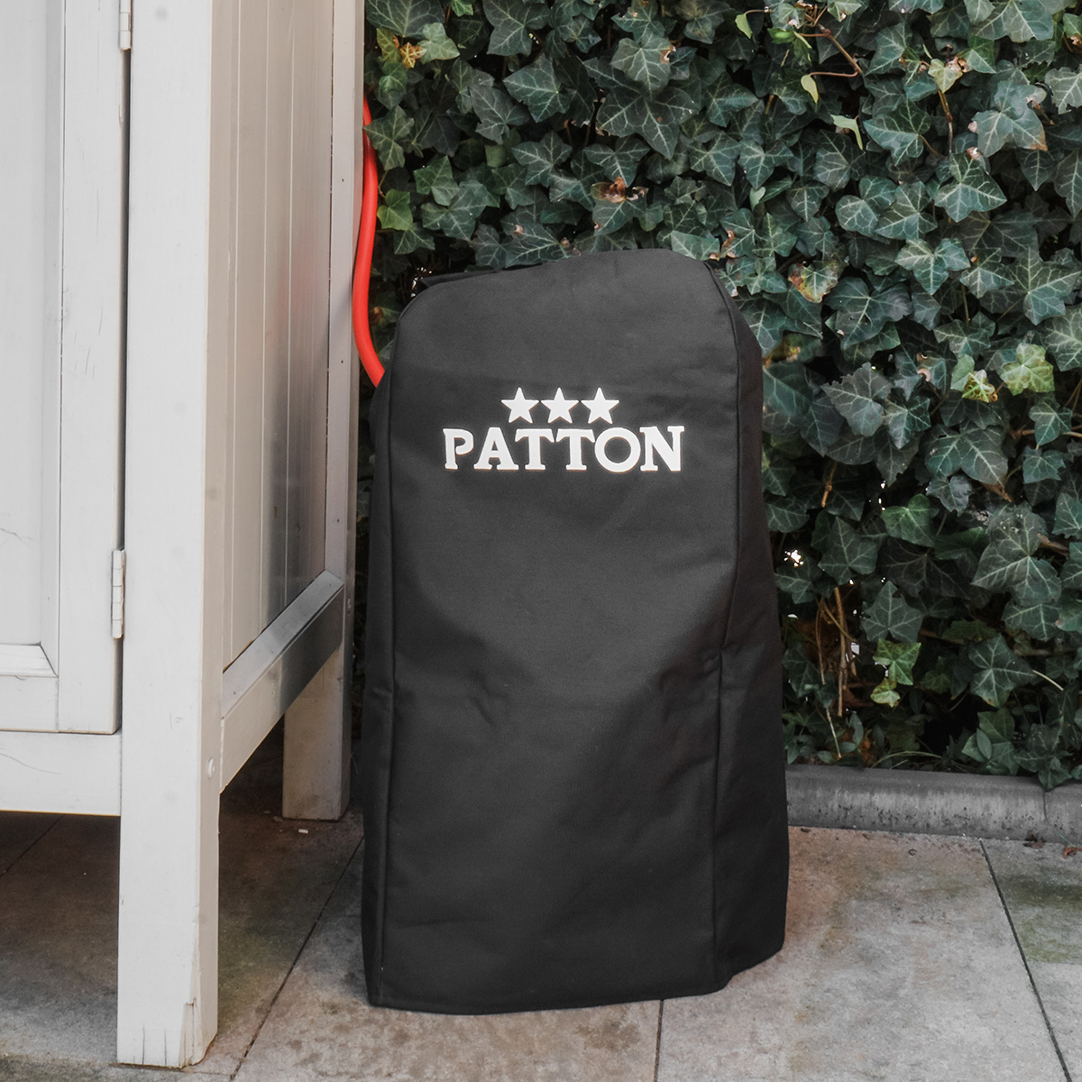 Een zwarte buitenhoes voor de grill met het witte woord 'PATTON' erop, in de buurt van klimop.