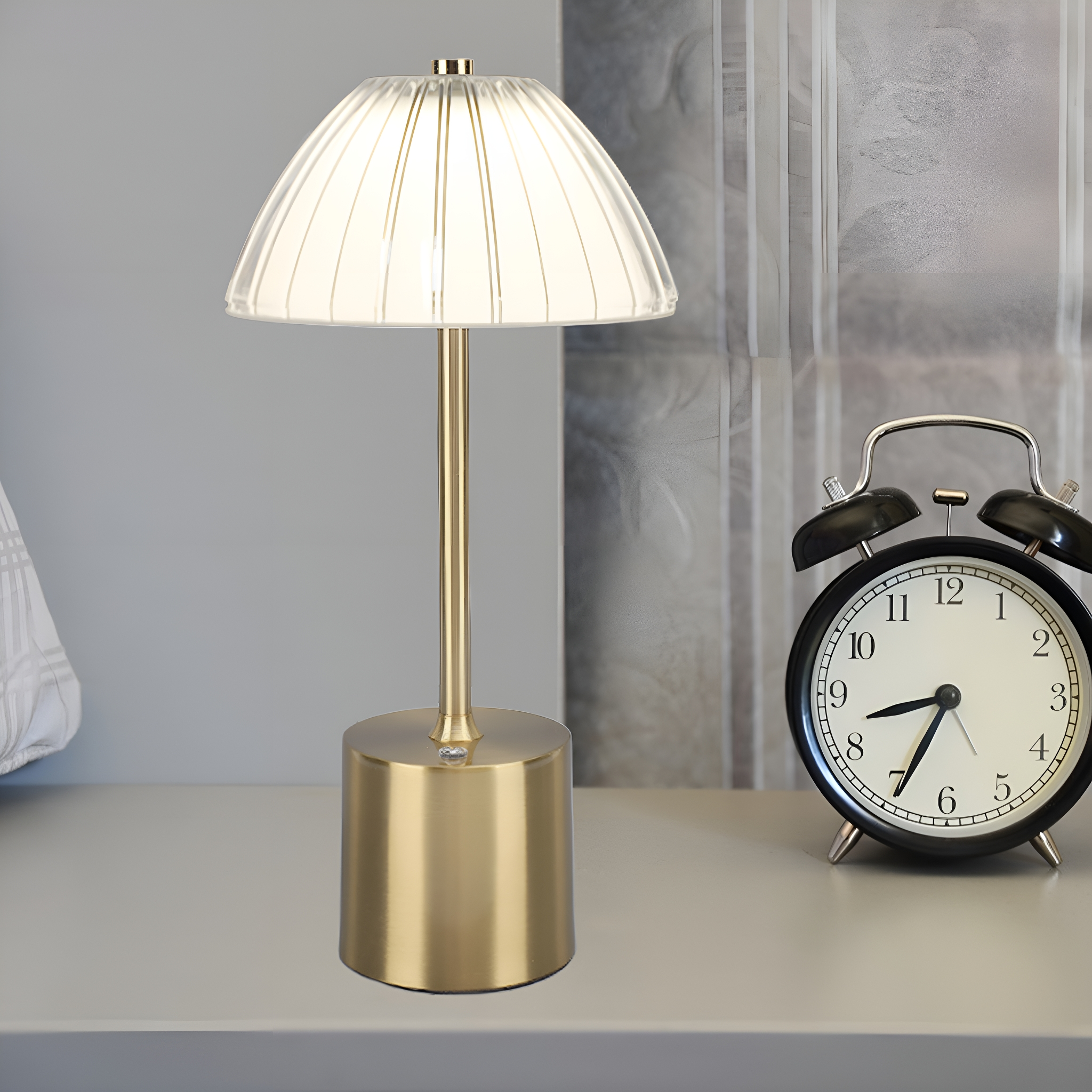 Eine leuchtende goldene Lampe mit weißem Schirm, neben einem schwarzen Wecker auf weißer Oberfläche.